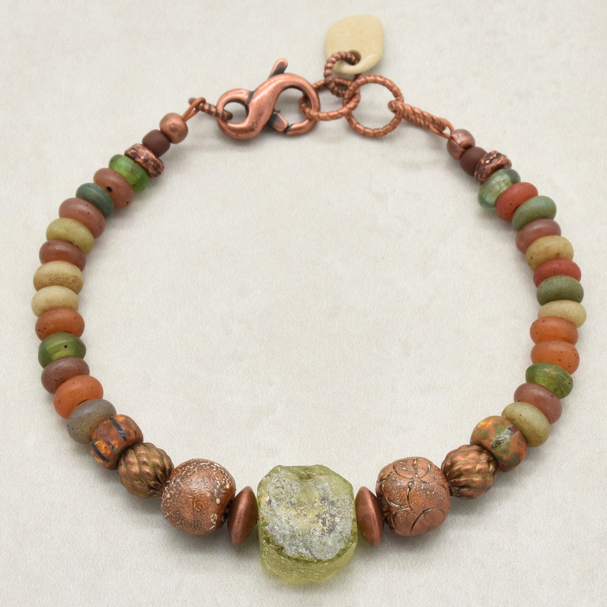 A handmade beaded bracelet featuring a mix of earthy Indonesian glass beads in terracotta, olive green, ochre, and brown, accented by textured copper beads, a central raw green ancient Roman glass focal, artisan ceramic beads, and a copper clasp on a light beige background - made with love in Colorado by Ruby Acorn. 