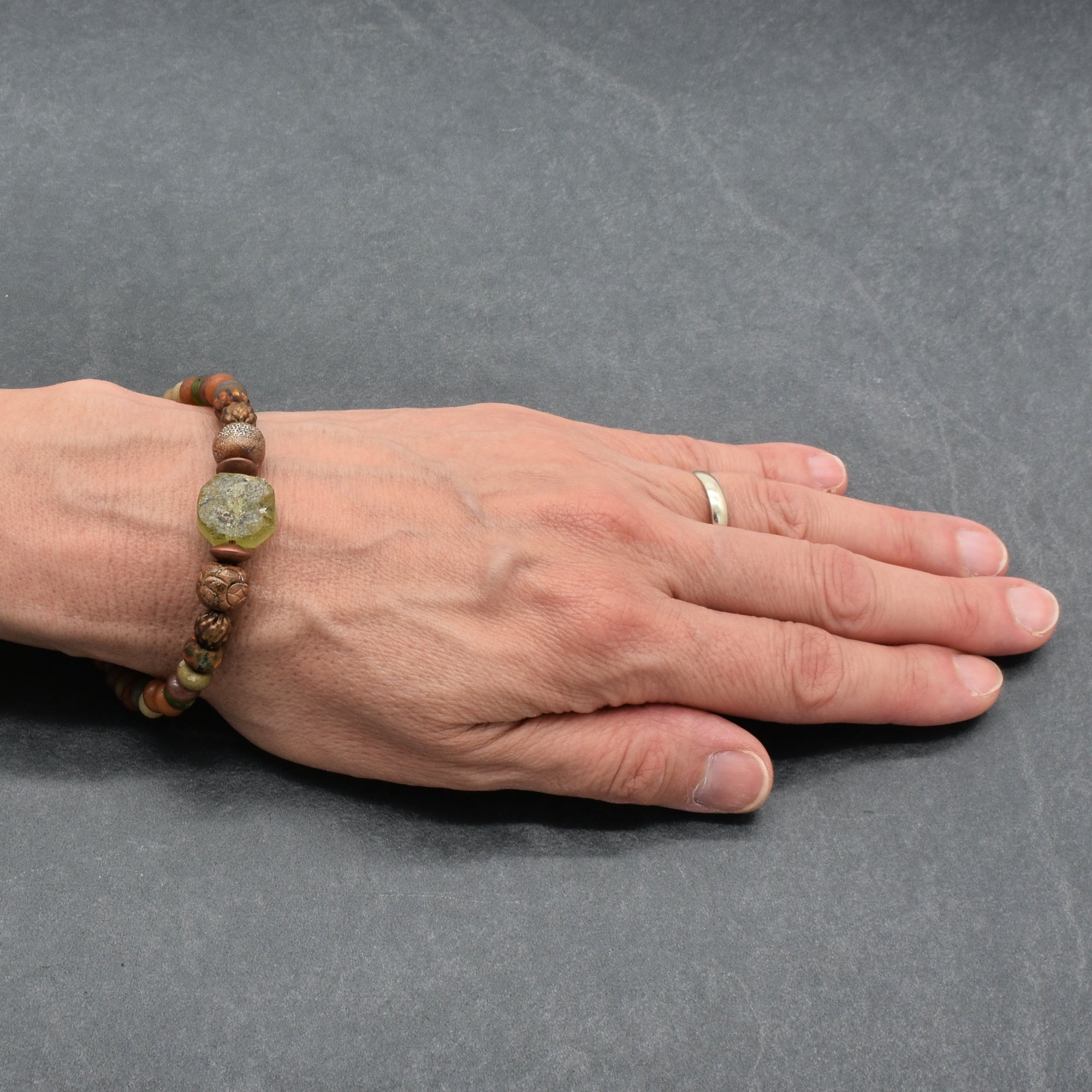 A close-up of a hand resting on a dark gray surface, wearing a beaded bracelet made of brown and green beads - made with love in Colorado by Ruby Acorn. 