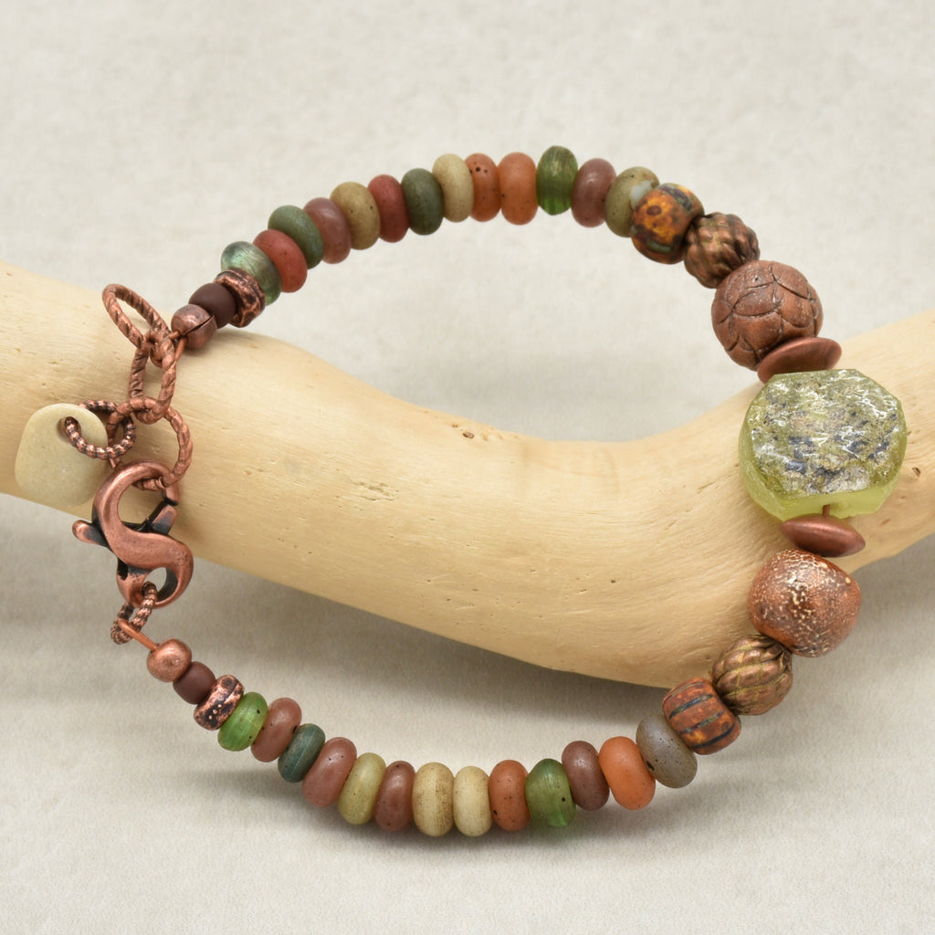 A handmade beaded bracelet with a mix of earthy brown, green, orange, and gray Indonesian glass beads, featuring a large textured green ancient Roman glass focal, artisan ceramic and a copper-toned lobster clasp, displayed on a curved light wood stand - made with love in Colorado by Ruby Acorn. 
