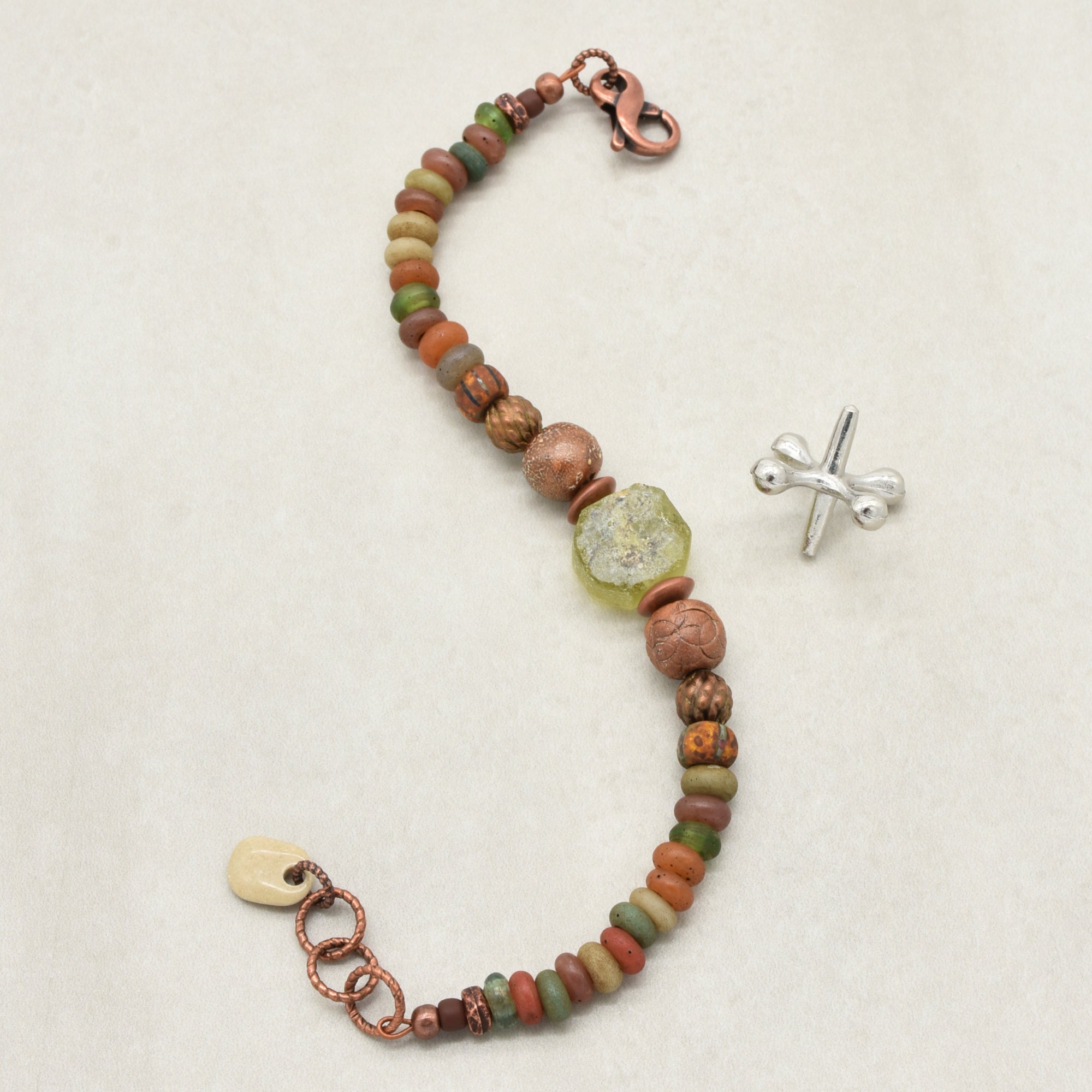 A handmade beaded bracelet with a mix of earthy brown, green, and terracotta Indonesian glass beads, a textured green ancient Roman glass centerpiece, a copper-toned lobster clasp, and a copper ring extender on a light beige background - made with love in Colorado by Ruby Acorn. 