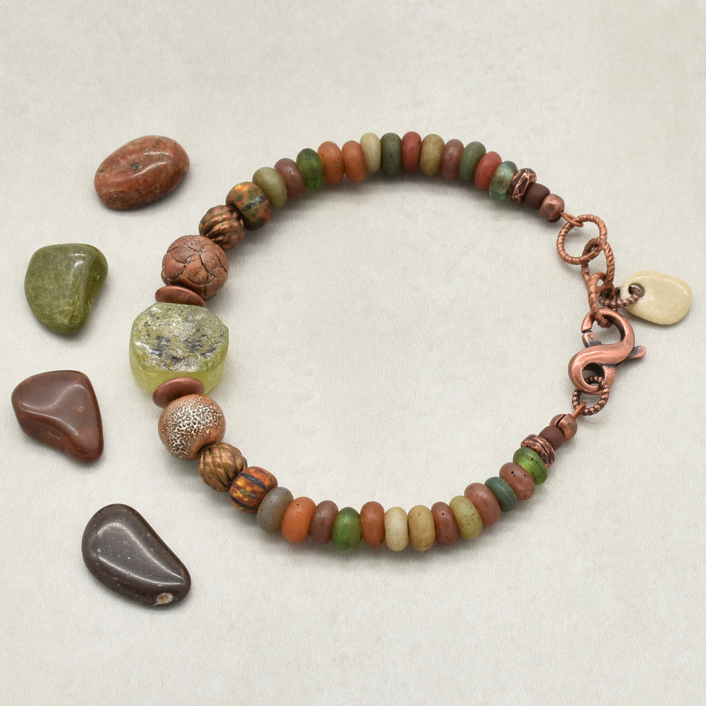A handmade beaded bracelet with a copper-toned clasp, featuring a mix of earthy Indonesian glass beads in brown, green, orange, and cream, a green ancient Roman glass focal, and artisan ceramic, displayed alongside five matching polished tumbled stones on a light beige background - made with love in Colorado by Ruby Acorn. 