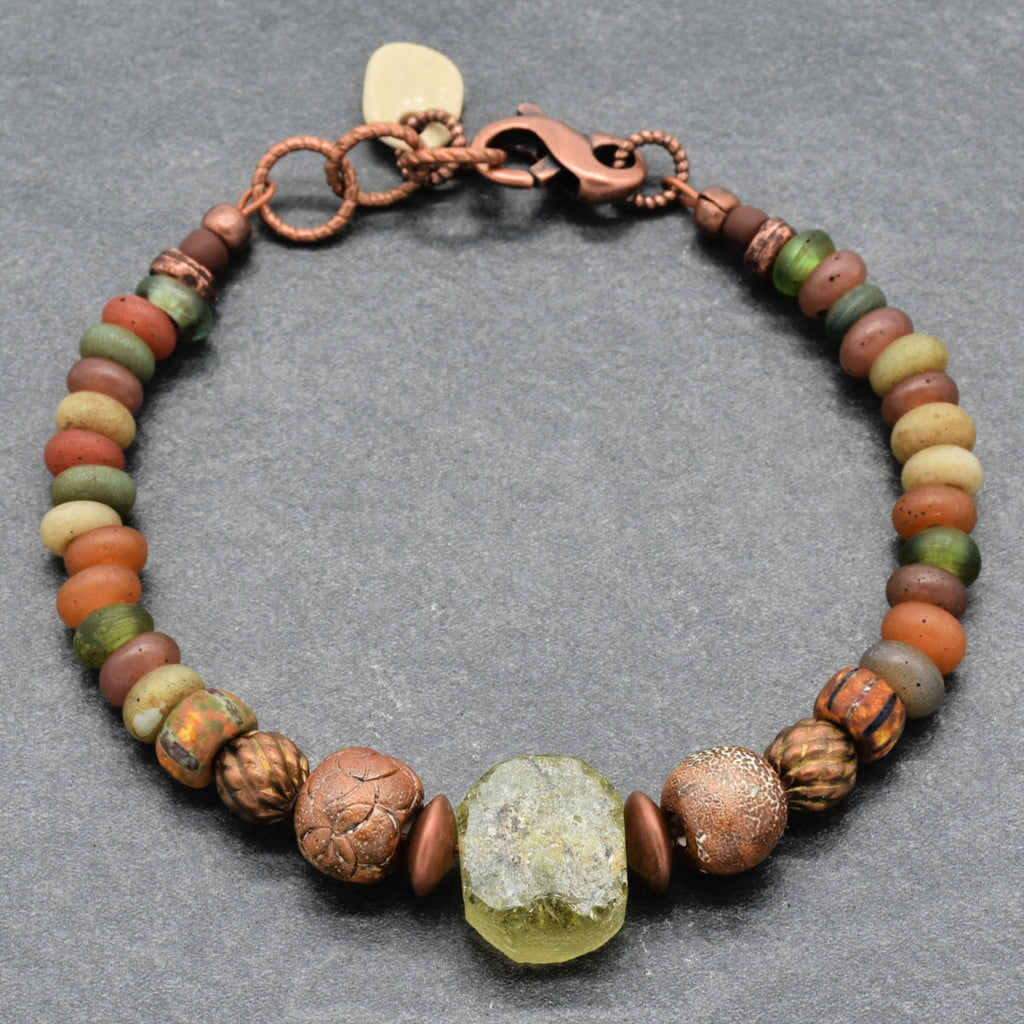 A handmade beaded bracelet featuring a variety of earthy, textured Indonesian glass beads in brown, green, orange, and terracotta, with a central translucent green ancient Roman glass focal, artisan ceramic beads, and a copper-toned clasp, displayed on a dark gray textured background - made with love in Colorado by Ruby Acorn. 
