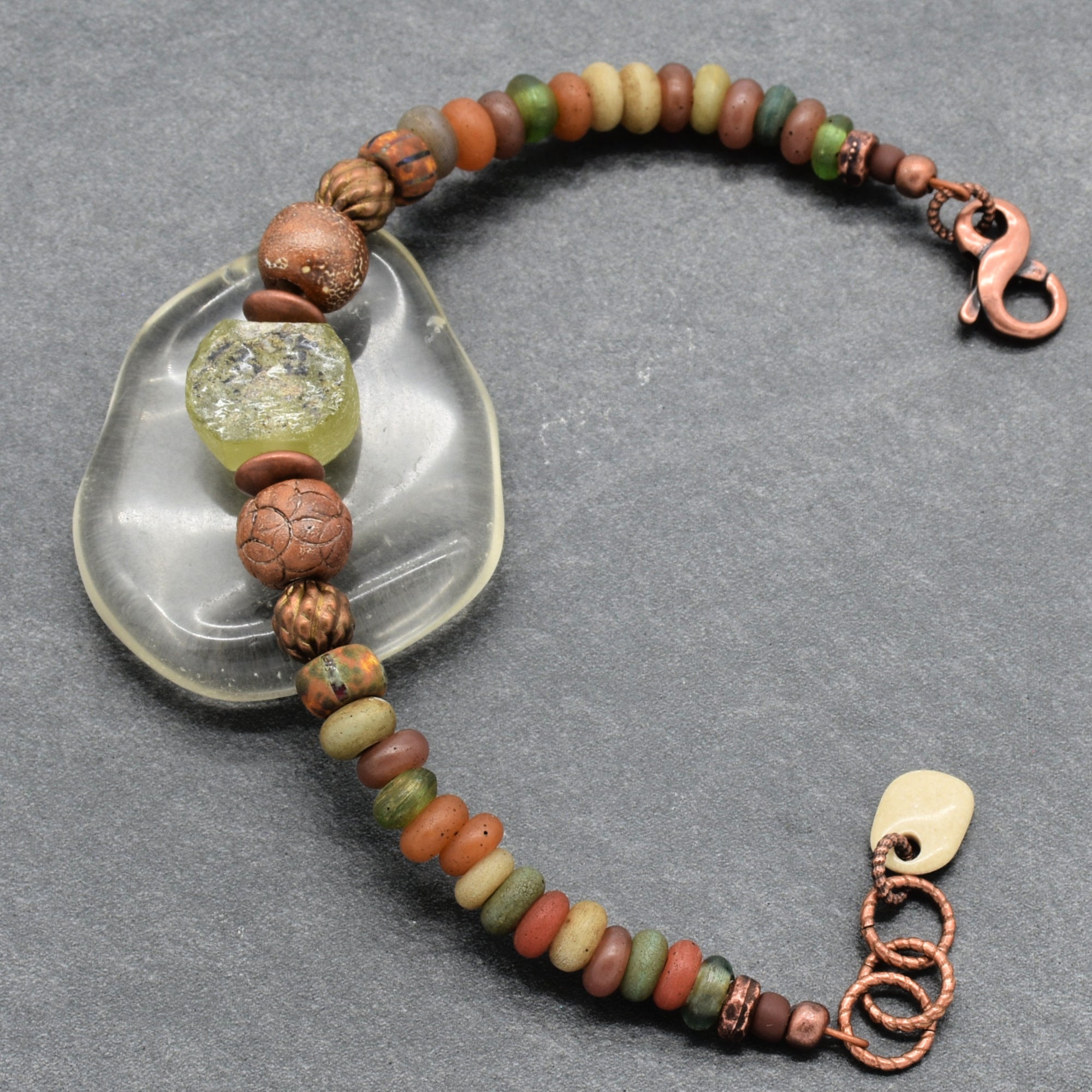 A handmade beaded bracelet with a mix of earthy brown, green, orange, and beige Indonesian glass beads, featuring a green ancient Roman glass focal, artisan ceramic, and a copper-toned lobster clasp, displayed on a gray background with a clear stone prop - made with love in Colorado by Ruby Acorn. 