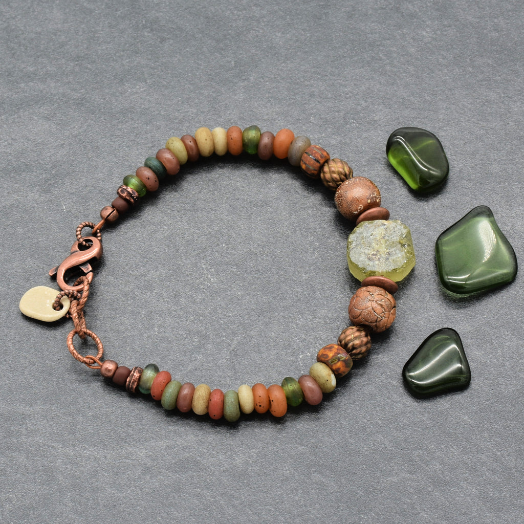 A handmade beaded bracelet with a copper clasp, displayed on a textured gray background alongside three polished dark green stones. The bracelet features a mix of earthy Indonesian glass beads in brown, orange, green, and beige, including a prominent raw green ancient Roman glass focal, artisan ceramic, and a small white drilled beach stone charm - made with love in Colorado by Ruby Acorn. 