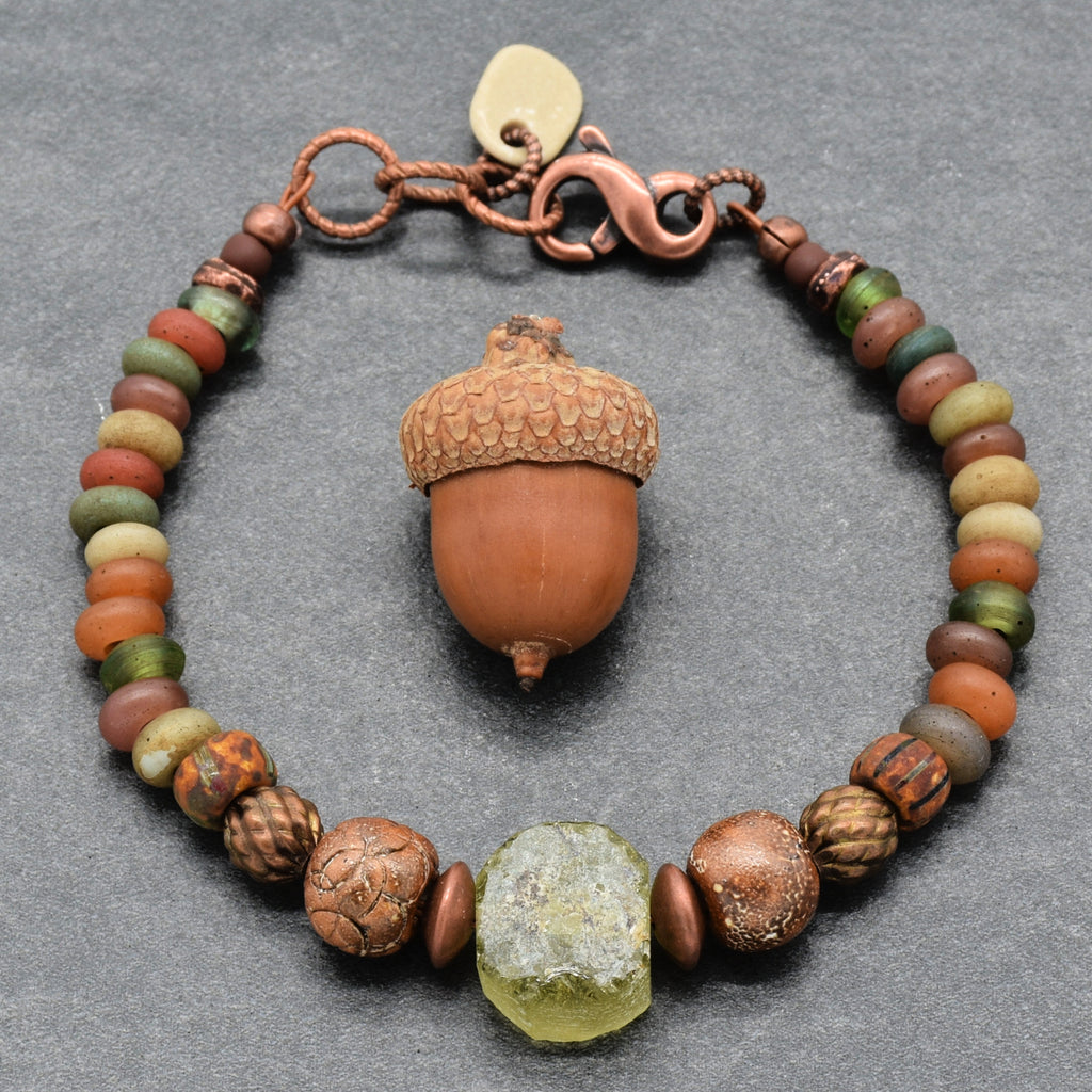 A handmade beaded bracelet featuring a textured green ancient Roman glass focal, and various earthy Indonesian glass beads in brown, green, and orange, arranged on a dark gray background - made with love in Colorado by Ruby Acorn. 