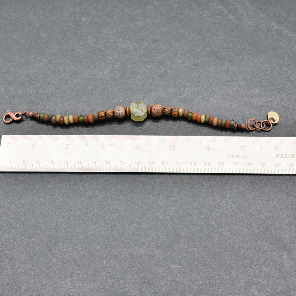 A handmade beaded bracelet with copper-toned metal clasp and chain, featuring various earthy green, brown, and red beads, including a central carved green stone, shown next to a white ruler on a dark gray background - made with love in Colorado by Ruby Acorn.