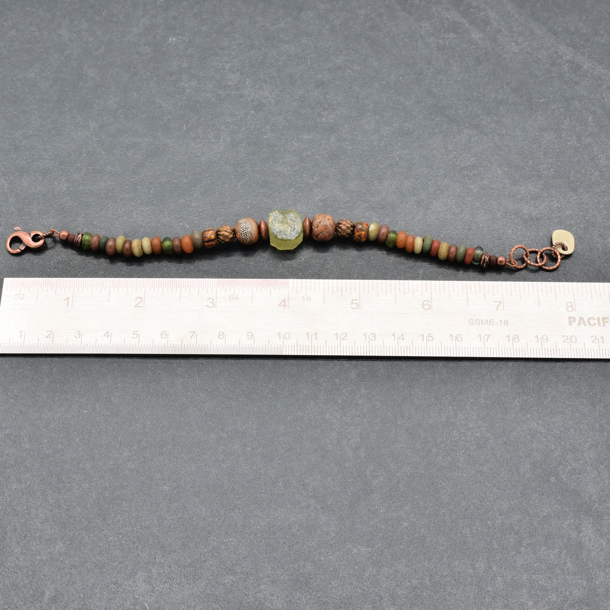 A handmade beaded bracelet with copper-toned metal clasp and chain, featuring various earthy green, brown, and red beads, including a central carved green stone, shown next to a white ruler on a dark gray background - made with love in Colorado by Ruby Acorn.