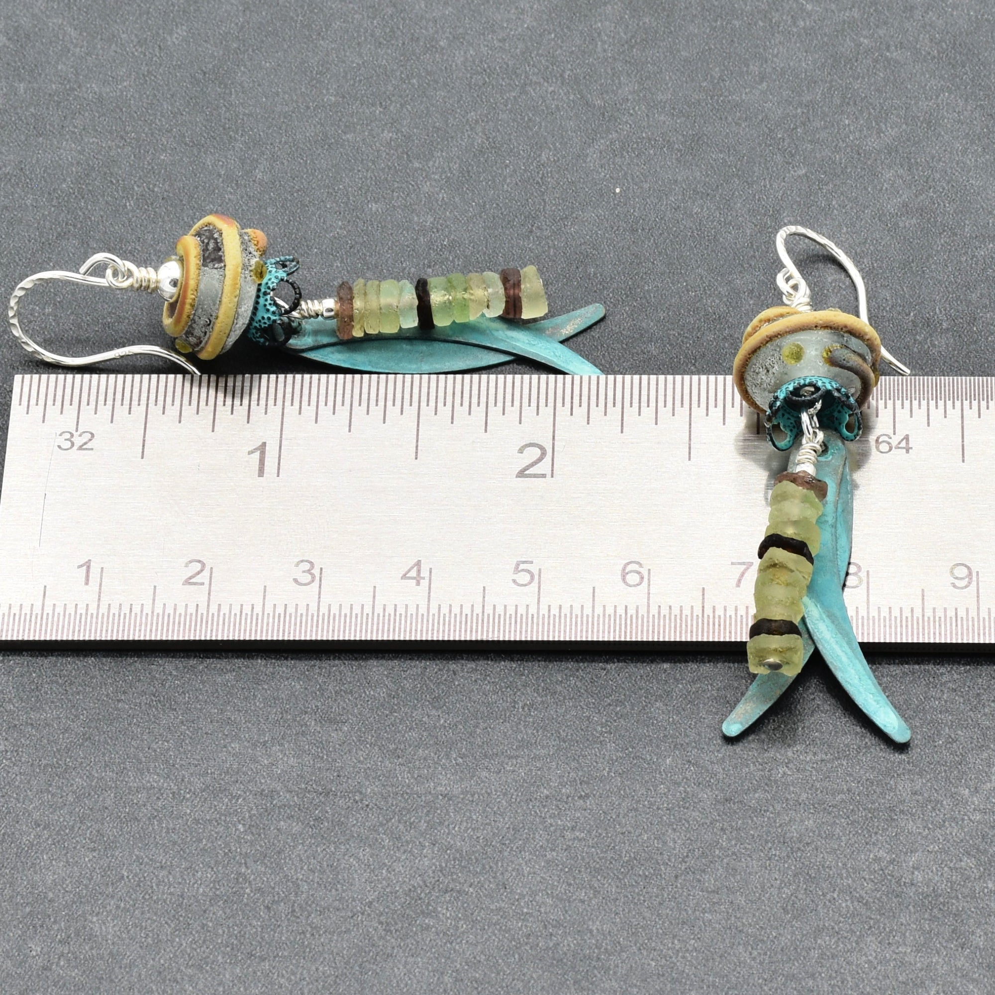 A pair of handmade dangle earrings displayed by a ruler for scale. The earrings feature silver ear wires, artisan lampwork glass, ancient Roman glass, and verdigris daggers - made with love in Colorado by Ruby Acorn.