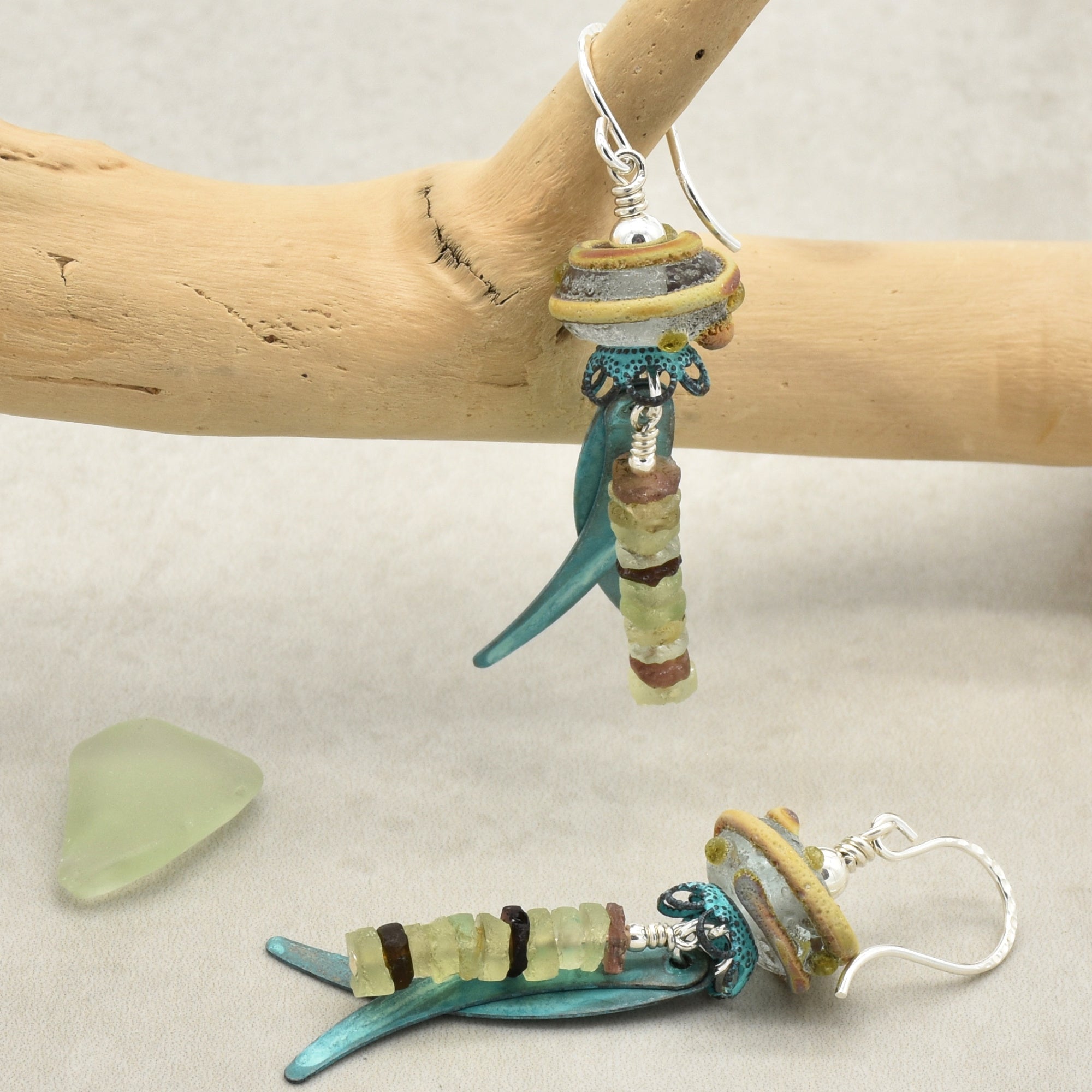 Handmade glass dangle earrings displayed on a branch, with one earring resting on a neutral surface beside a small piece of sea glass. The earrings feature ancient Roman glass, artisan lampwork glass, verdigris daggers, and silver ear wires - made with love in Colorado by Ruby Acorn.