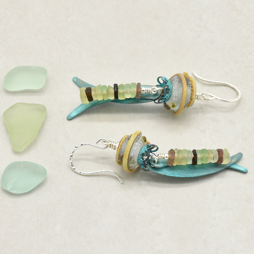 A pair of handmade dangle earrings made from ancient Roman glass, artisan lampwork glass, verdigris daggers and silver ear wires, displayed on a light beige background next to three pieces of light green sea glass - made with love in Colorado by Ruby Acorn.