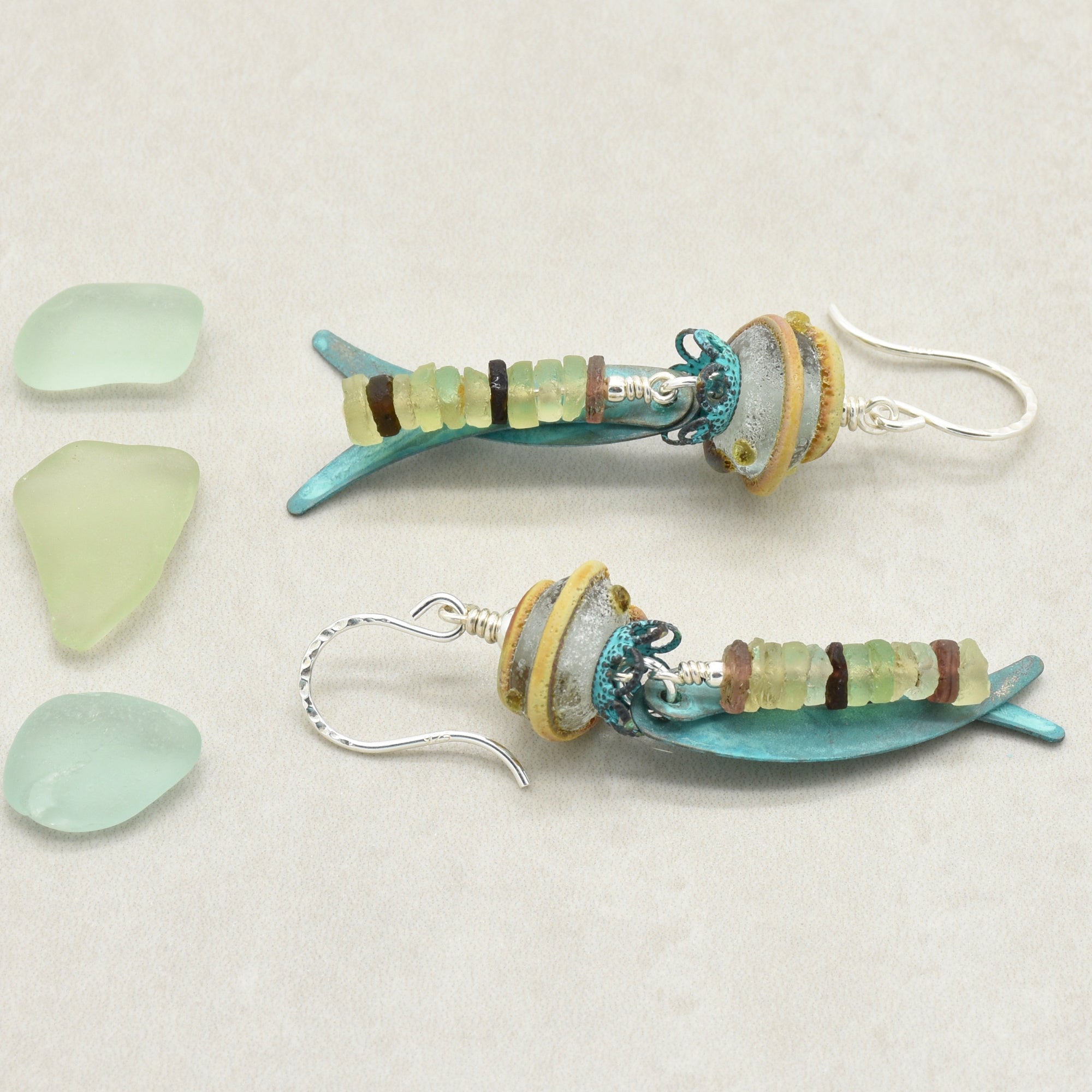 A pair of handmade dangle earrings made from ancient Roman glass, artisan lampwork glass, verdigris daggers and silver ear wires, displayed on a light beige background next to three pieces of light green sea glass - made with love in Colorado by Ruby Acorn.
