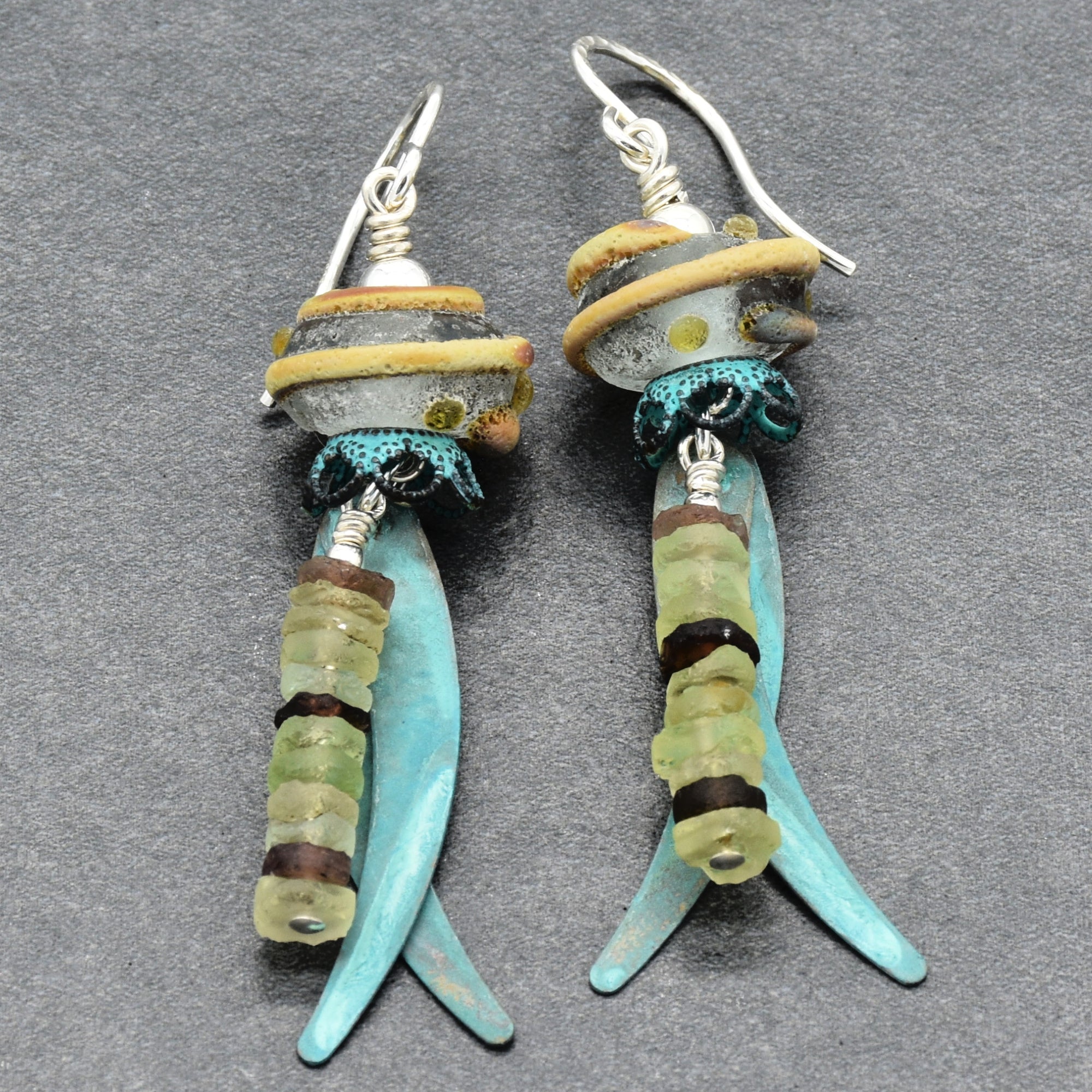 A pair of handmade dangle earrings featuring ancient Roman glass, artisan lampwork glass, verdigris daggers,  silver ear wires, displayed on a dark gray textured background - made with love in Colorado by Ruby Acorn.