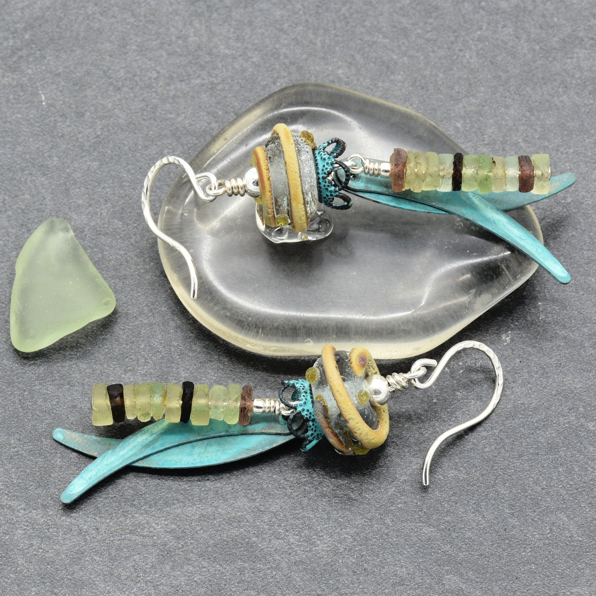 Handmade glass dangle earrings displayed on a dark gray textured surface, with one earring resting on a clear glass dish and the other beside a small piece of sea glass. The earrings feature ancient Roman glass, artisan lampwork glass, verdigris daggers, and silver ear wires - made with love in Colorado by Ruby Acorn.