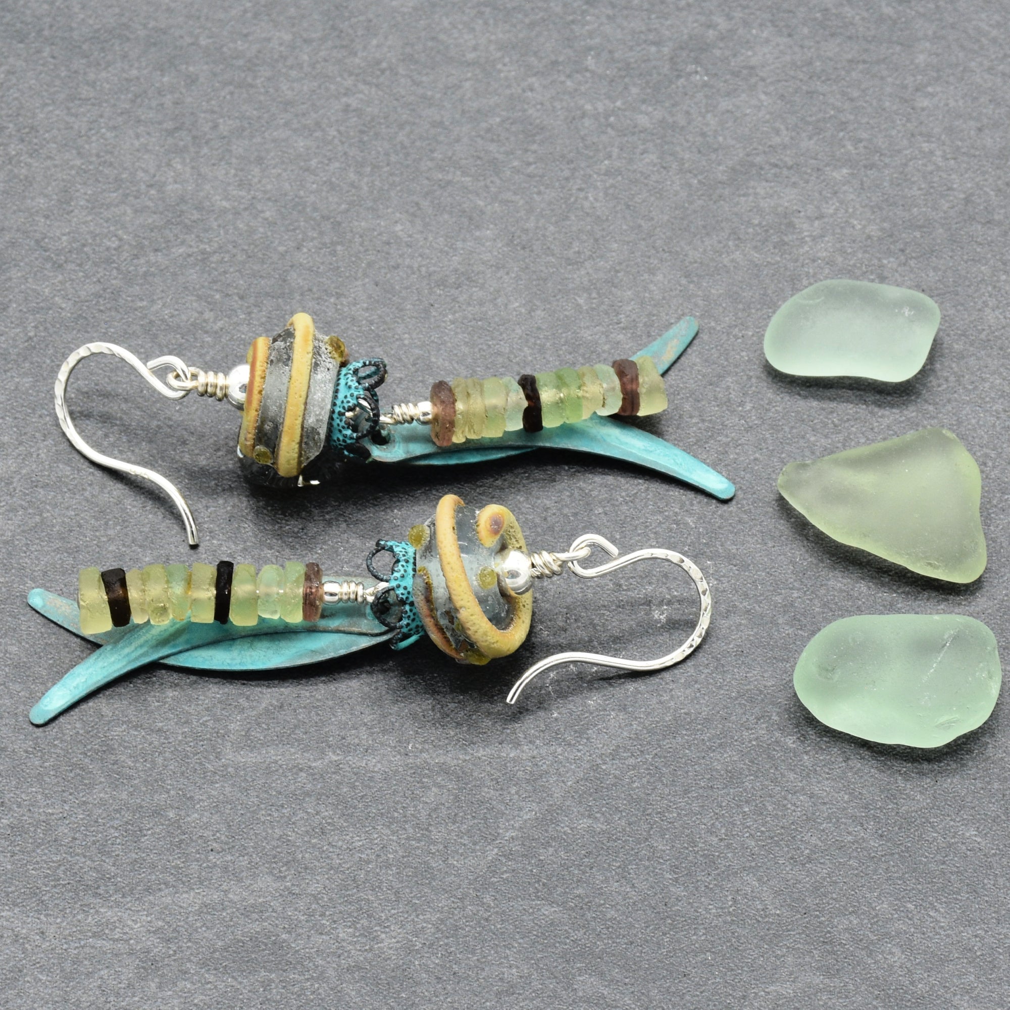 A pair of handmade dangle earrings displayed on a textured gray background next to three loose sea glass pieces. The earrings feature ancient Roman glass, artisan lampwork glass, verdigris daggers, and silver ear wires - made with love in Colorado by Ruby Acorn.
