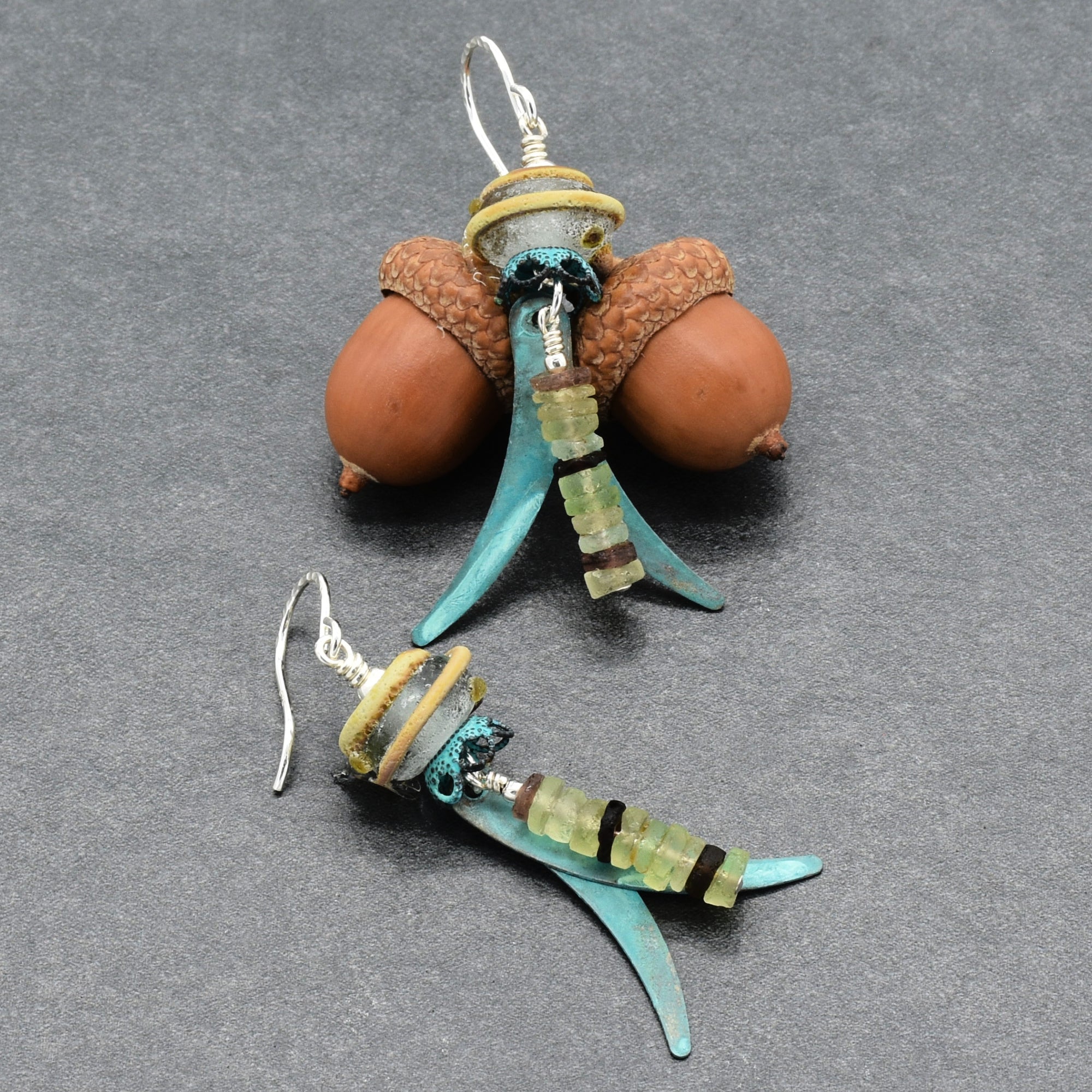 A pair of handmade dangle earrings made from ancient Roman glass, artisan lampwork glass, verdigris daggers and silver ear wires, displayed on an acorn on a gray background - made with love in Colorado by Ruby Acorn.