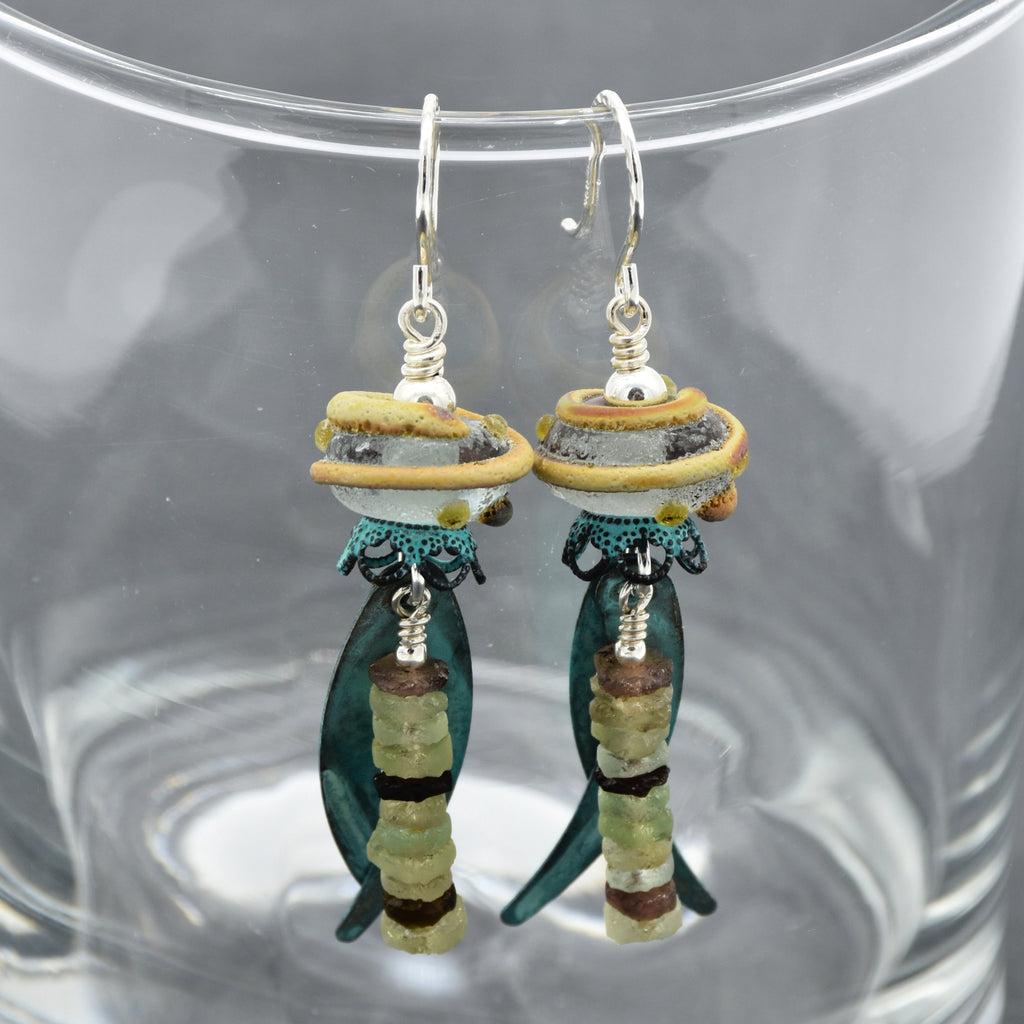 A pair of handmade dangle earrings displayed inside clear glass. The earrings feature silver ear wires, artisan lampwork glass, ancient Roman glass, and verdigris daggers - made with love in Colorado by Ruby Acorn.
