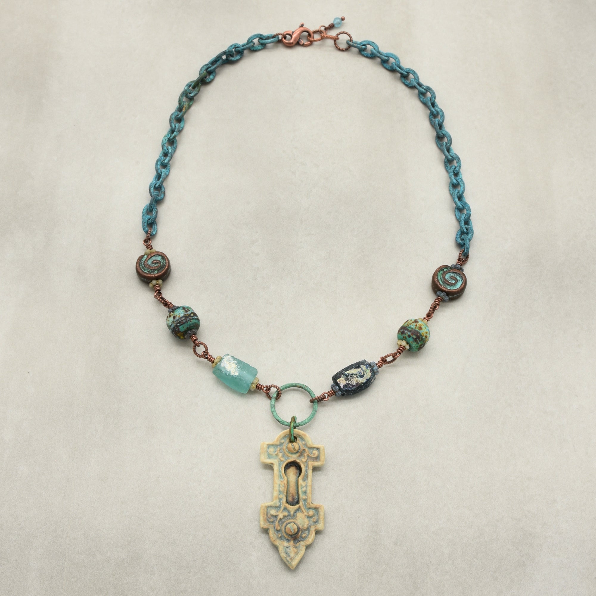 A bohemian-style beaded necklace featuring a verdigris chain, copper-toned wire, ancient Roman glass beads, and an artisan porcelain keyhole focal, displayed on a neutral beige background - made with love in Colorado by Ruby Acorn.