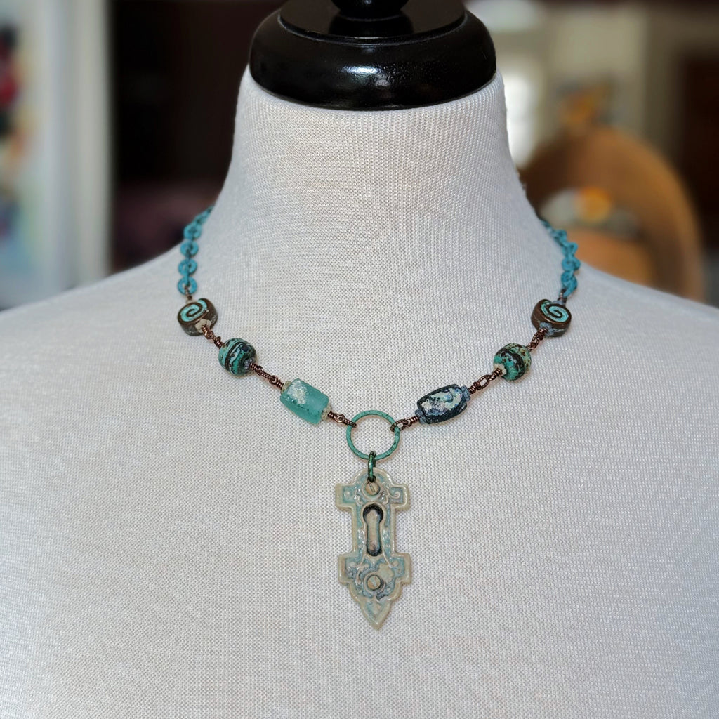 A wire-wrapped necklace featuring an artisan porcelain keyhole pendant, ancient Roman glass, and verdigris chain displayed on a cream fabric mannequin bust - made with love in Colorado by Ruby Acorn.