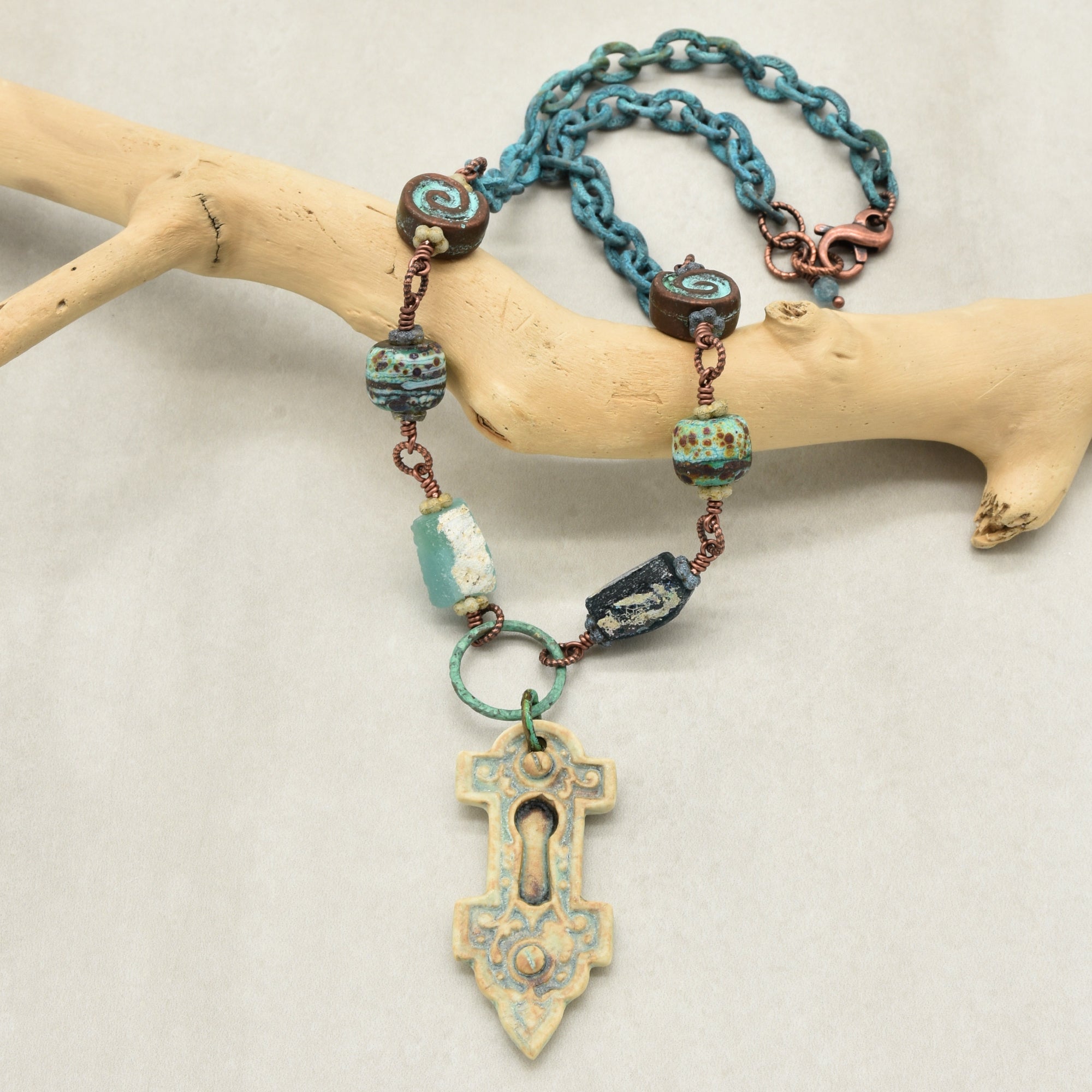 A bohemian-style keyhole pendant necklace with verdigris chain, and ancient Roman glass beads, displayed on a light wood branch against a neutral background - made with love in Colorado by Ruby Acorn.