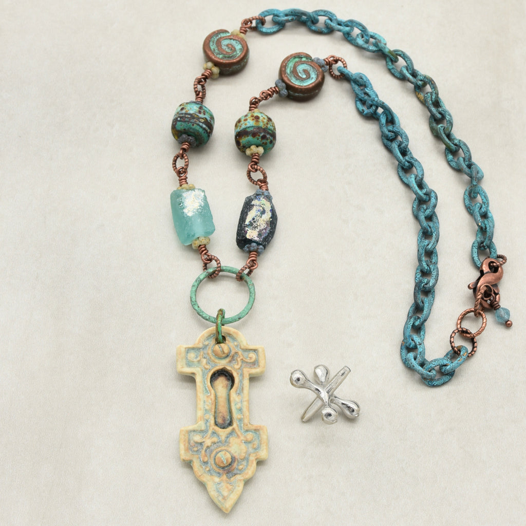 A handmade necklace with an artisan porcelain keyhole pendant, ancient Roman glass beads, and verdigris chain, displayed on a light beige background - made with love in Colorado by Ruby Acorn.