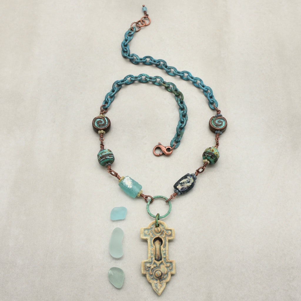 A bohemian-style wire-wrapped necklace with verdigris chain, Mykonos ammonite beads, artisan lampwork glass beads, ancient Roman glass, and an artisan porcelain keyhole pendant, displayed on a neutral beige background - made with love in Colorado by Ruby Acorn.
