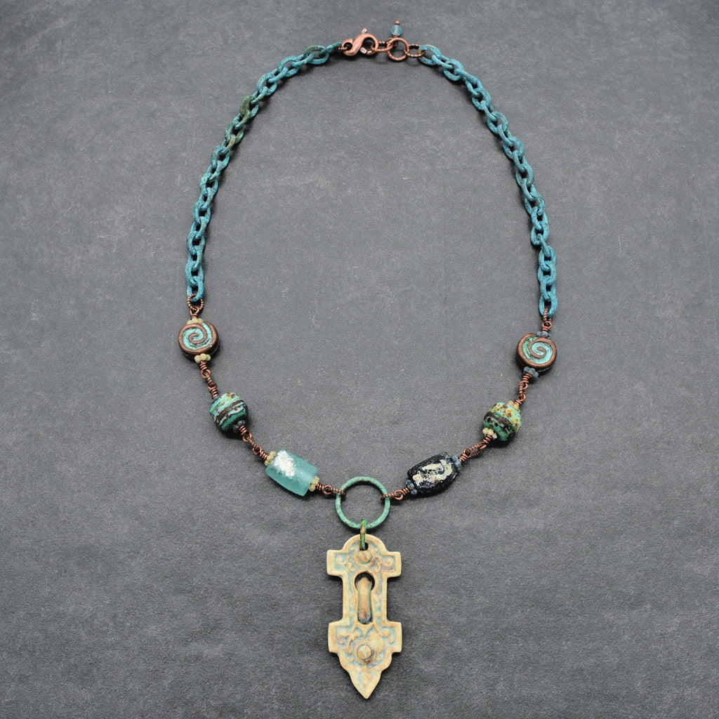 An artisanal verdigris chain necklace with ancient Roman glass beads and an artisan porcelain keyhole pendant, displayed on a textured gray background - made with love in Colorado by Ruby Acorn.