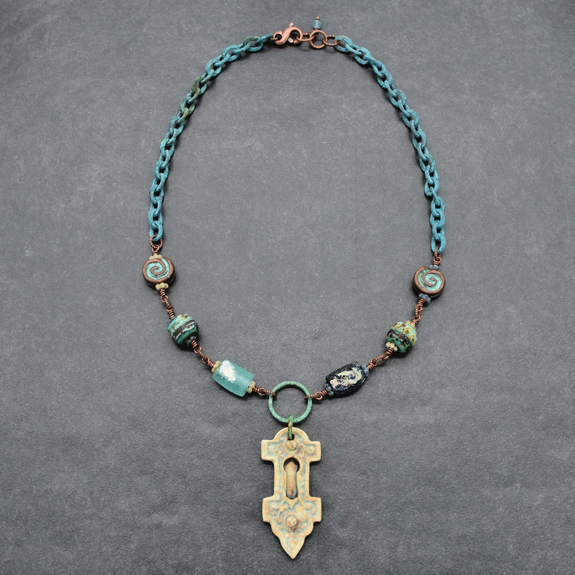 An artisanal verdigris chain necklace with ancient Roman glass beads and an artisan porcelain keyhole pendant, displayed on a textured gray background - made with love in Colorado by Ruby Acorn.