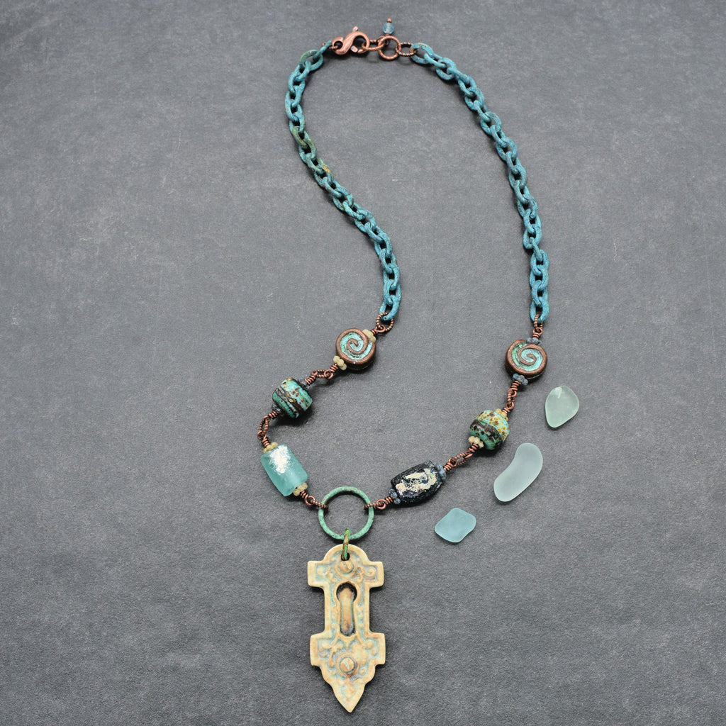 A handmade wire-wrapped necklace with ancient Roman glass, an artisan porcelain keyhole focal, artisan lampwork glass, Mykonos ammonite beads, and verdigris chain, displayed on a gray background - made with love in Colorado by Ruby Acorn.