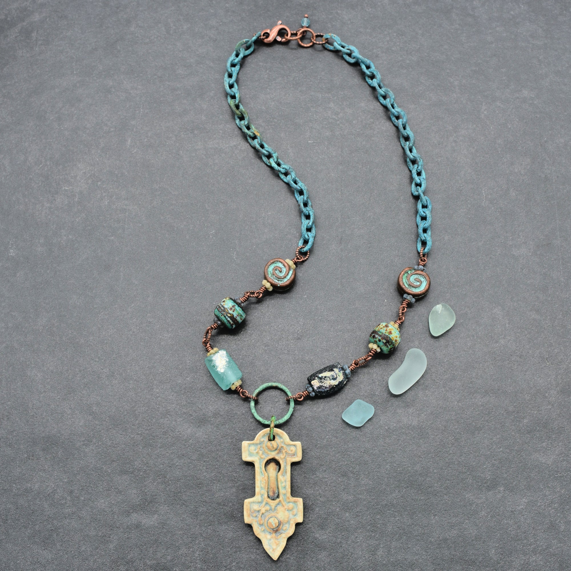 A handmade wire-wrapped necklace with ancient Roman glass, an artisan porcelain keyhole focal, artisan lampwork glass, Mykonos ammonite beads, and verdigris chain, displayed on a gray background - made with love in Colorado by Ruby Acorn.