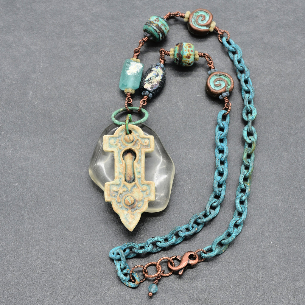 A handmade keyhole pendant necklace featuring ancient Roman glass beads, and verdigris chain, displayed on a dark gray textured background - made with love in Colorado by Ruby Acorn.