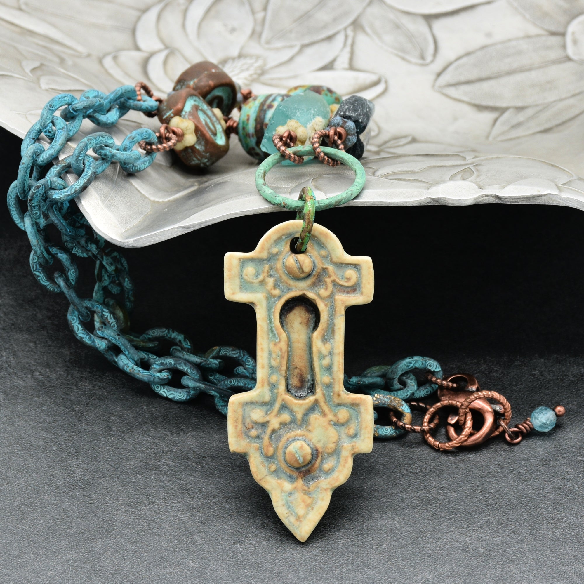 A handmade necklace with an artisan porcelain keyhole pendant, verdigris chain, and ancient Roman glass beads, displayed on a dark gray surface with a silver tray in the background - made with love in Colorado by Ruby Acorn.