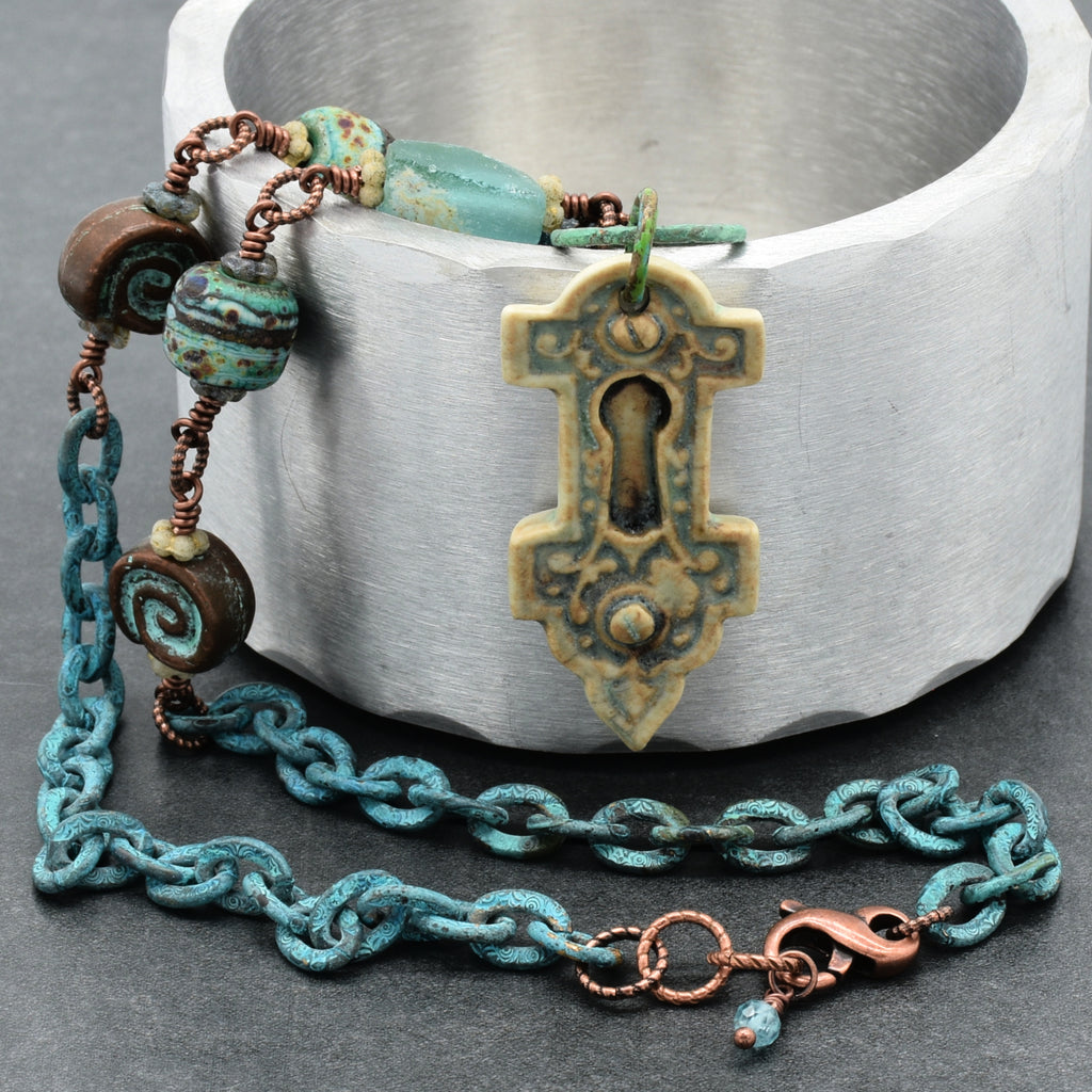 A closeup of a handmade necklace with an artisan porcelain keyhole pendant, ancient Roman glass, and verdigris chain on a silver display - made with love in Colorado by Ruby Acorn.