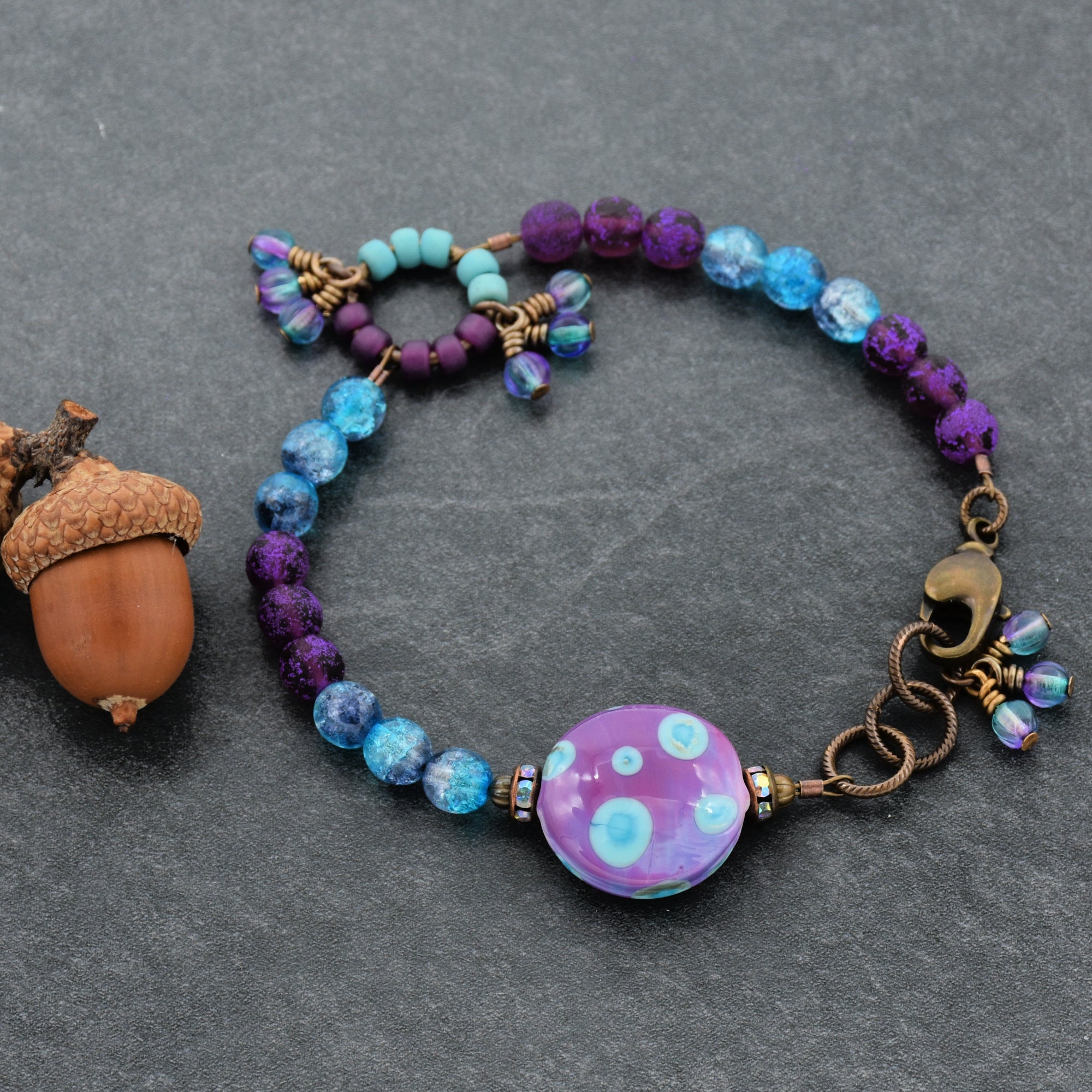 Colorful beaded bracelet with a polka dot bead on a gray surface next to an acorn.