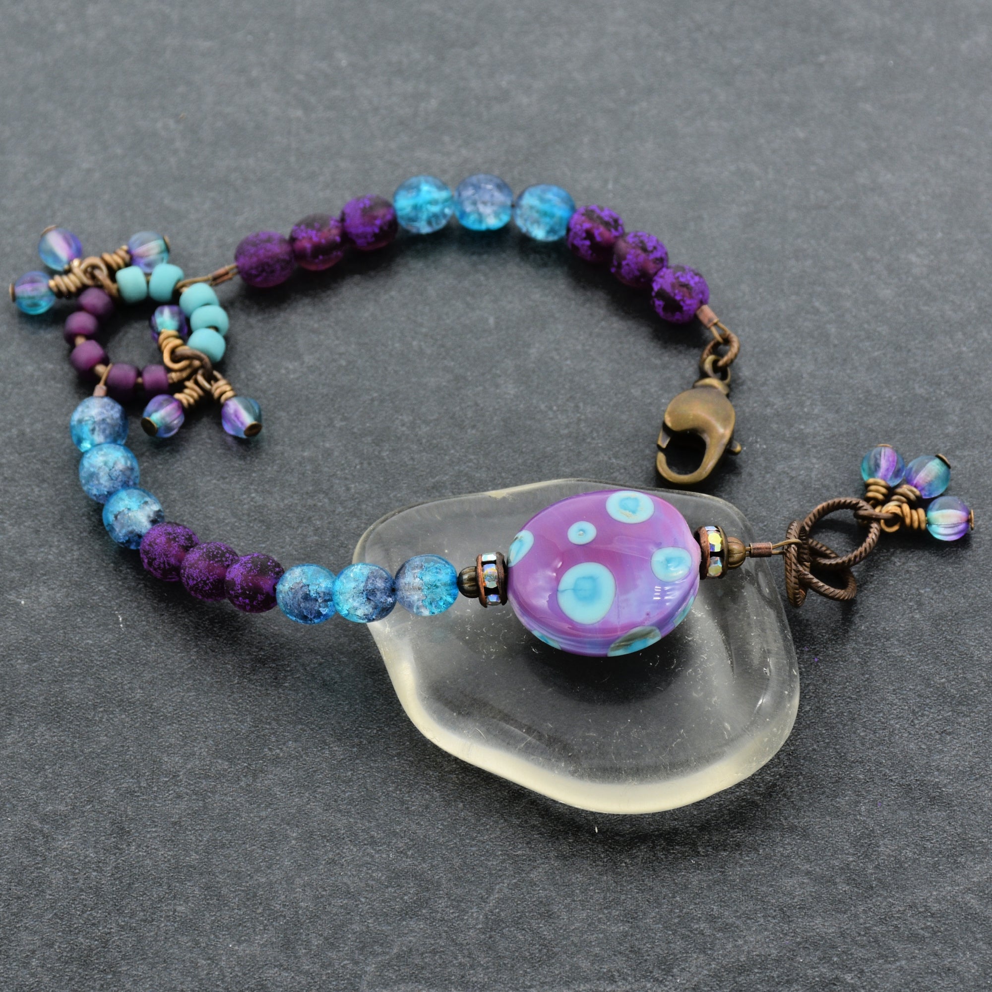 Beaded bracelet with a purple and blue polka dotted lampwork bead on a gray surface