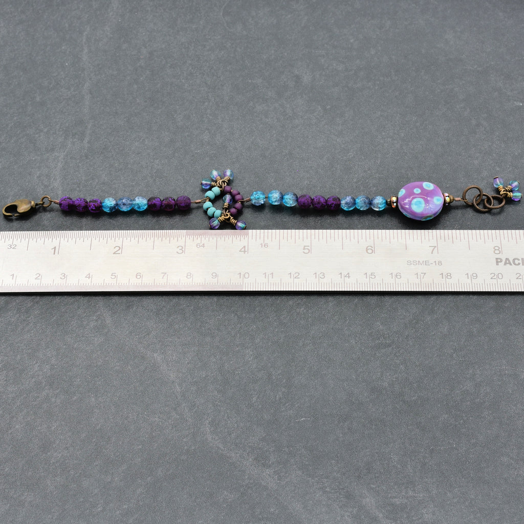 Beaded bracelet with a decorative purple and blue bead on a gray surface with a ruler for scale.
