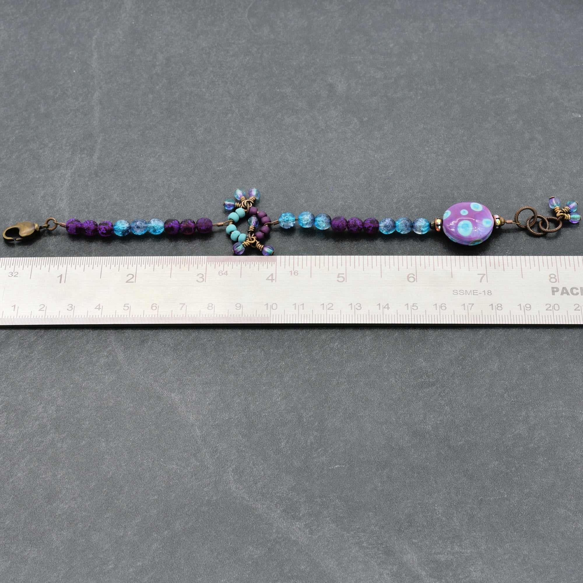 Beaded bracelet with a decorative purple and blue bead on a gray surface with a ruler for scale.