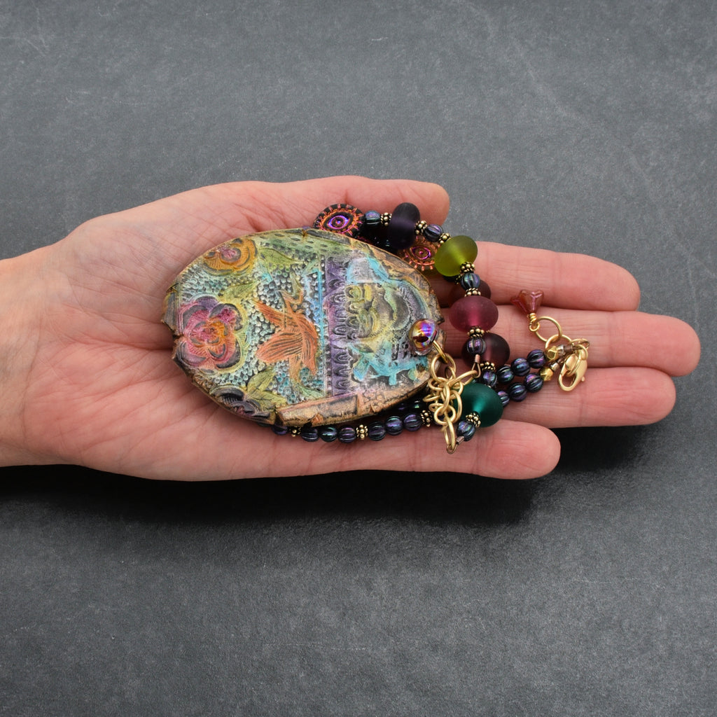Hand holding necklace with large decorative pendant with colorful patterns  against a gray background