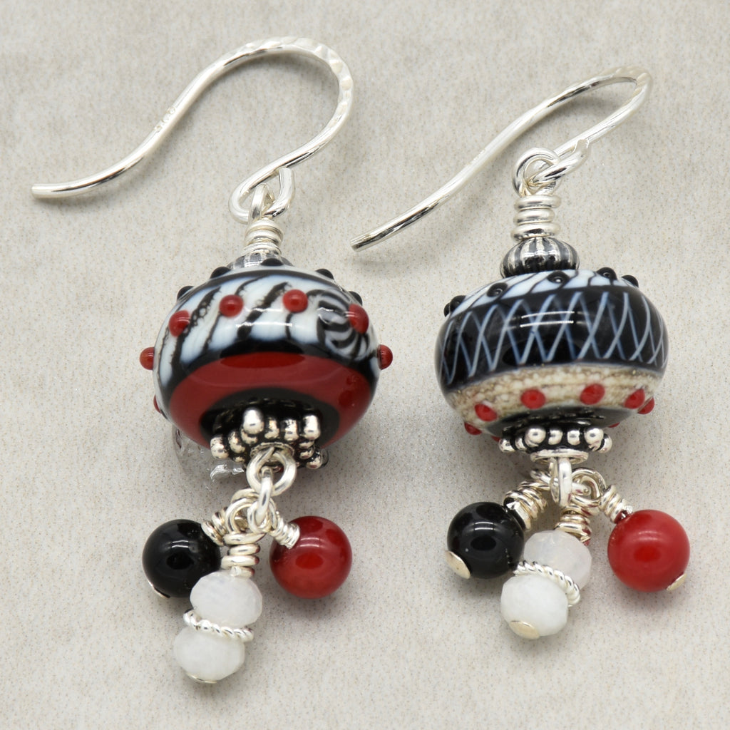 A pair of handmade dangle earrings with silver-toned fishhook wires, featuring patterned black and white glass beads with red accents, and dangling red, black, and white beads on a neutral gray background - made with love in Colorado by Ruby Acorn.