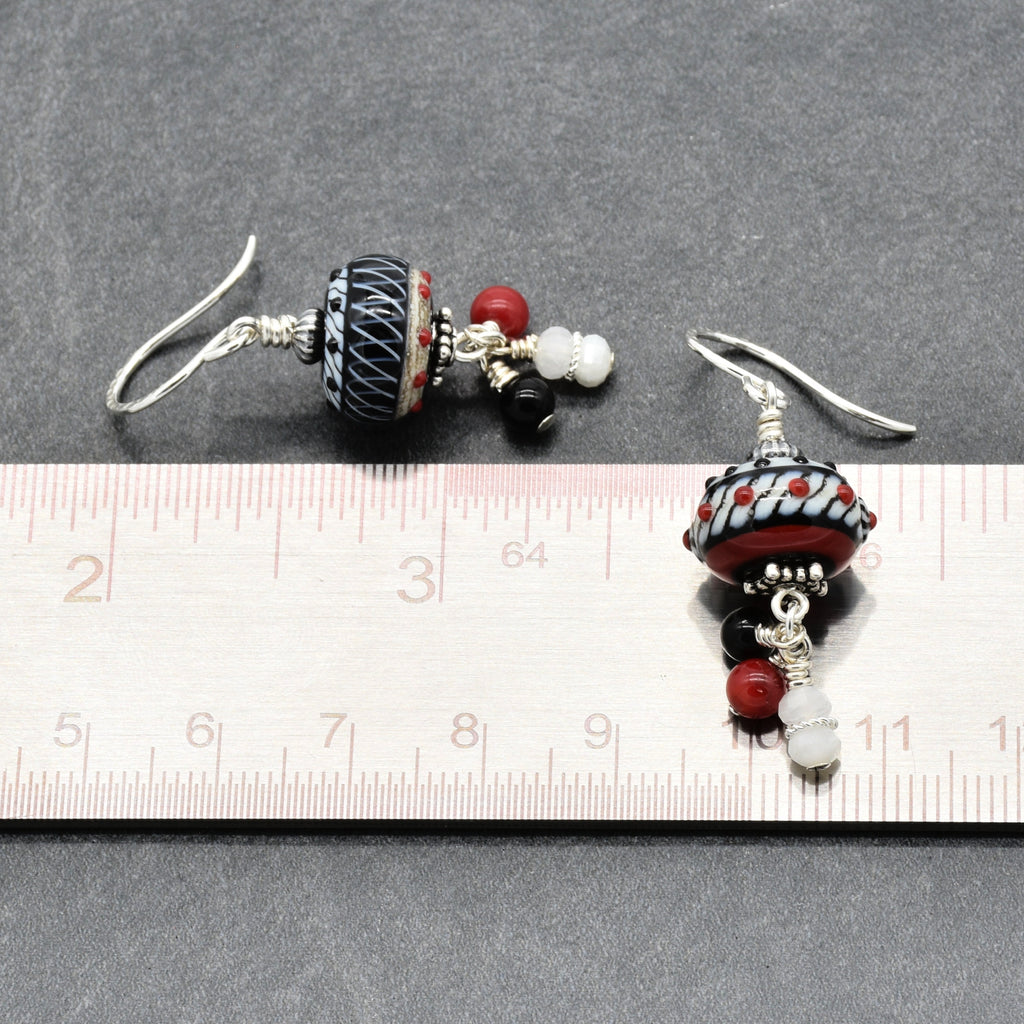 A pair of handmade dangle earrings displayed next to a metal ruler on a dark gray textured background. The earrings feature a large black and white patterned glass bead, red and white beads, and silver-toned wire hooks - made with love in Colorado by Ruby Acorn.