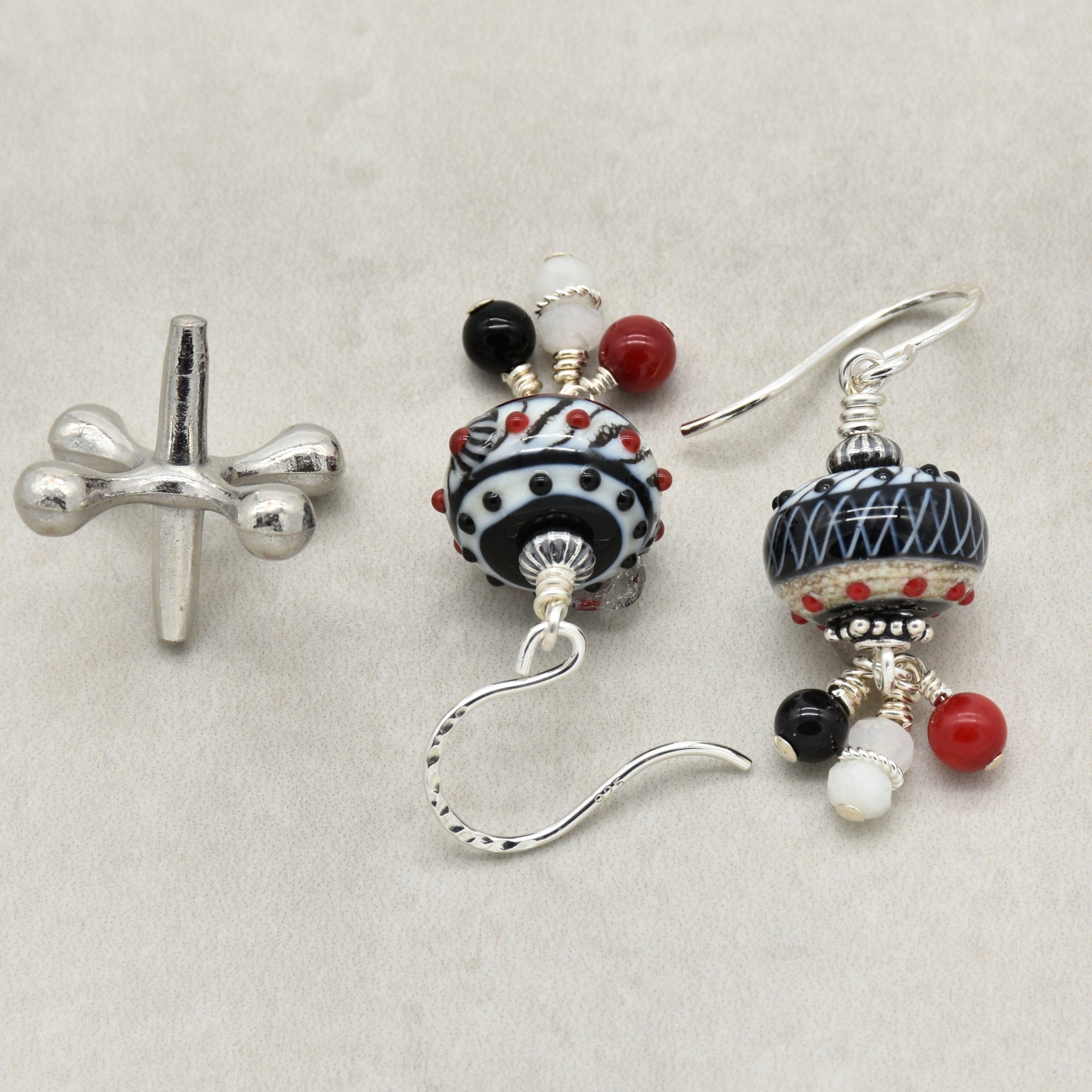 A pair of dangle earrings with patterned black and white beads, red bead accents, and small white beads on a textured light gray background - made with love in Colorado by Ruby Acorn.