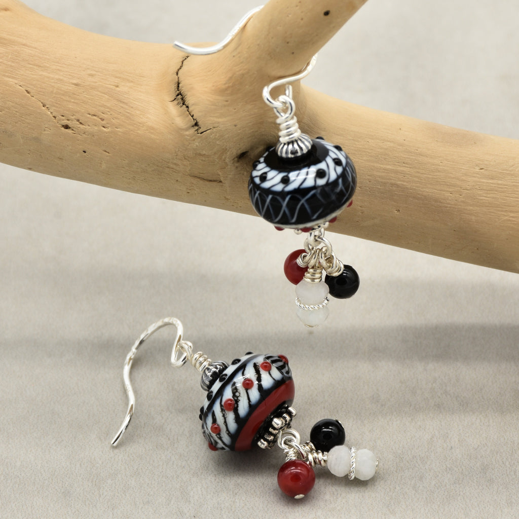 A pair of handmade dangle earrings with patterned black and white beads, red accents, and silver hooks, displayed on a light wood branch against a neutral gray background - made with love in Colorado by Ruby Acorn.