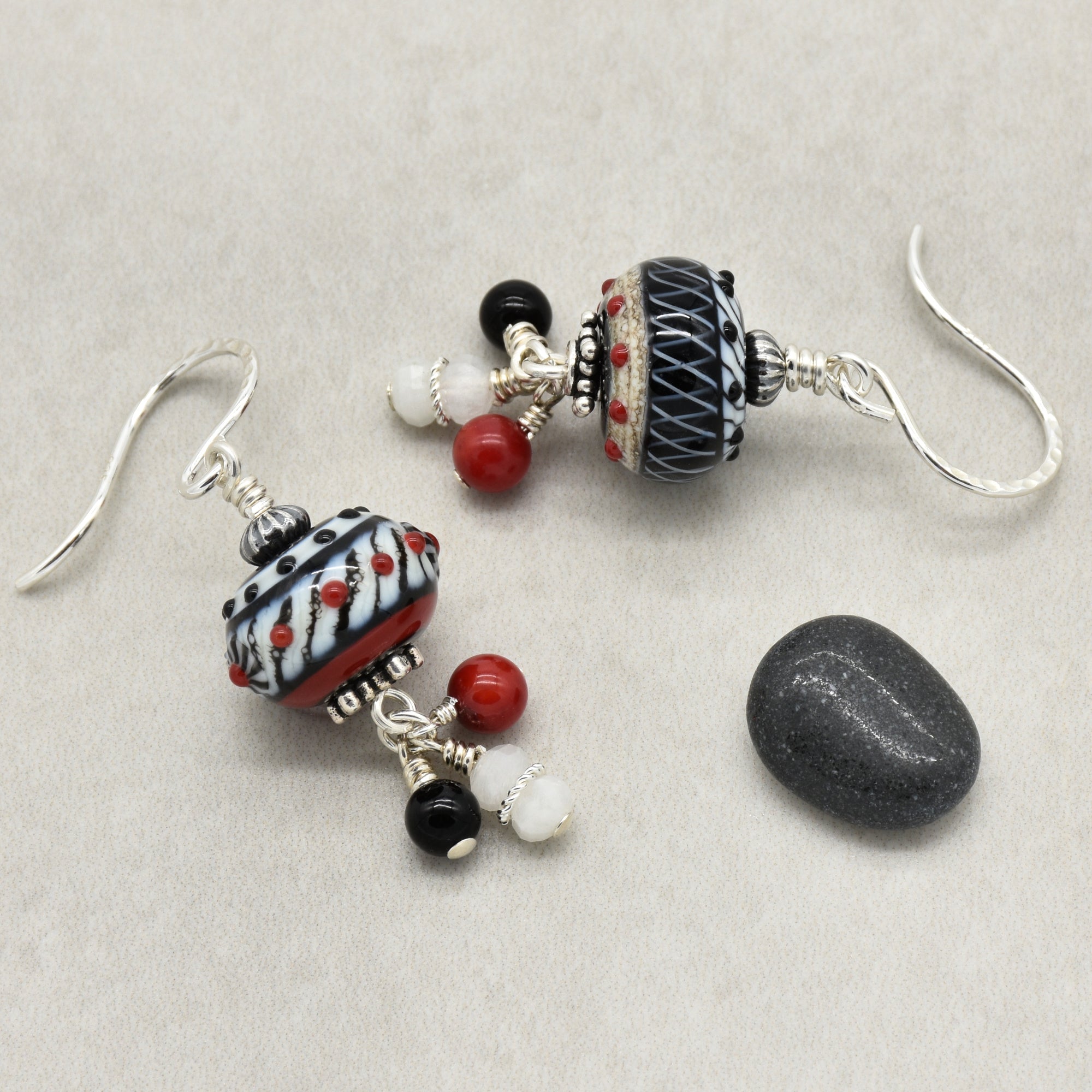 A pair of handmade dangle earrings with silver fishhook wires, featuring patterned black and white beads with tiny red accents, small red and white beads, and a dark gray stone charm, displayed on a neutral beige background - made with love in Colorado by Ruby Acorn.