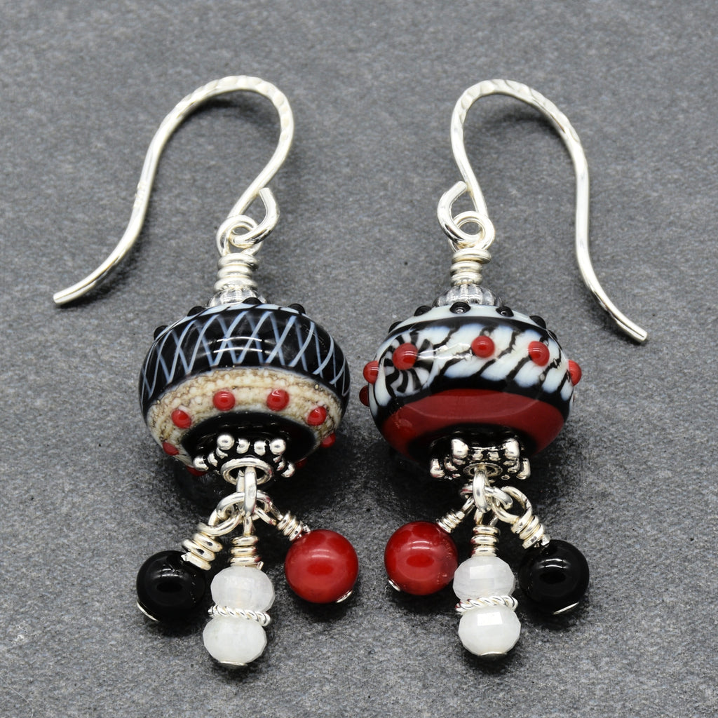 A pair of handmade dangle earrings with silver-toned French hook wires, featuring patterned black and white glass beads with red accents, and dangling black, red, and white beads on a textured gray background - made with love in Colorado by Ruby Acorn.