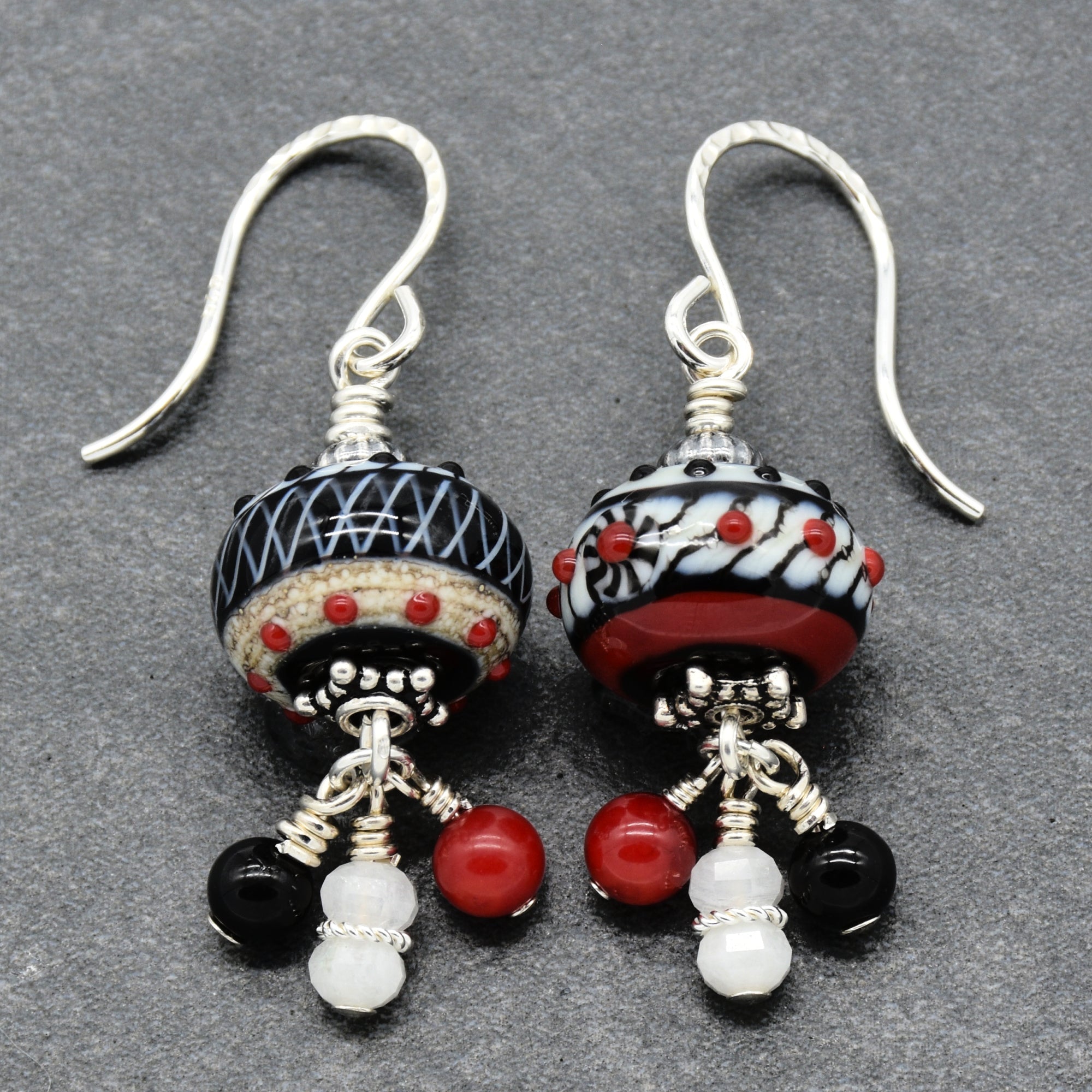 A pair of handmade dangle earrings with silver-toned French hook wires, featuring patterned black and white glass beads with red accents, and dangling black, red, and white beads on a textured gray background - made with love in Colorado by Ruby Acorn.