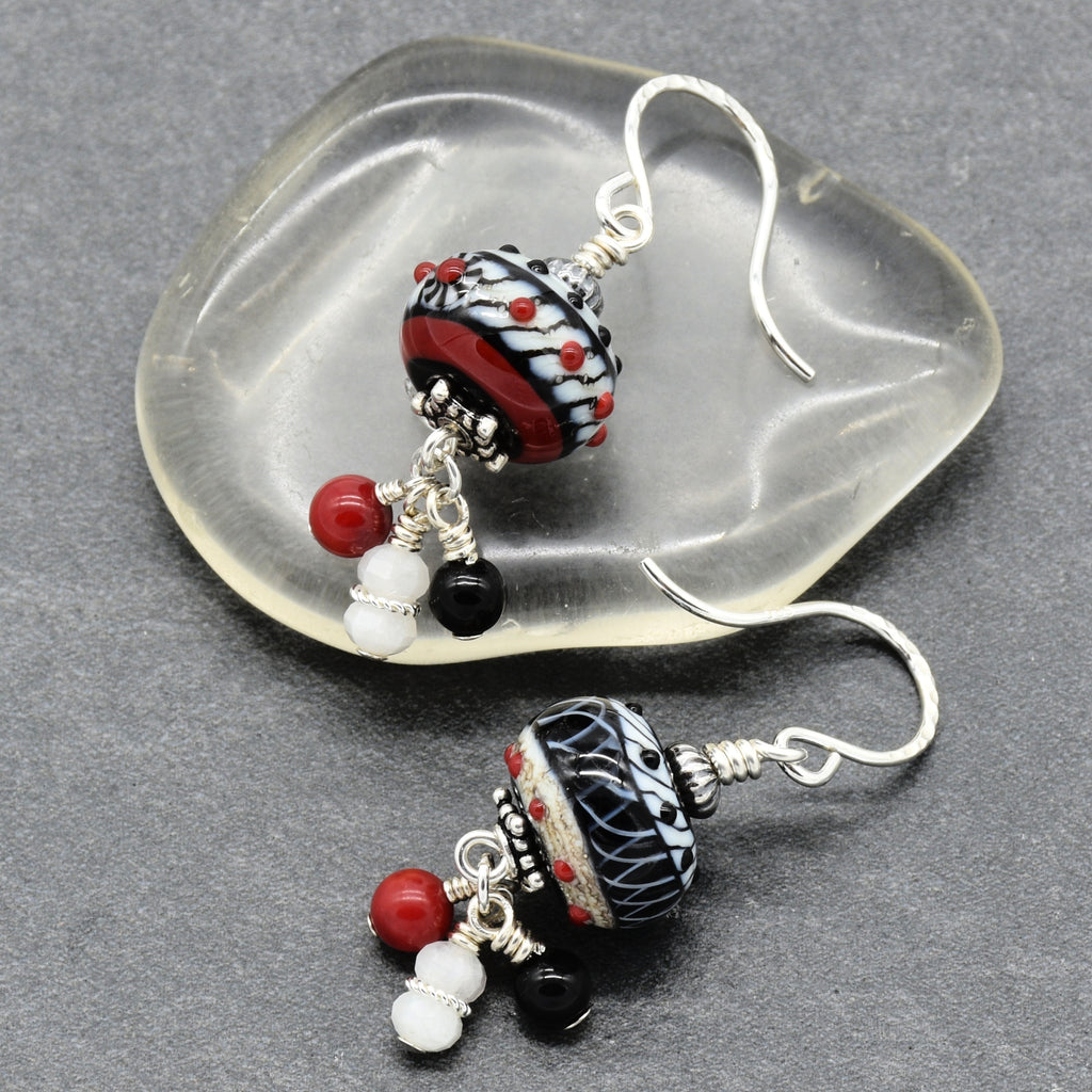 A pair of handmade lampwork dangle earrings displayed on a clear, organic-shaped glass dish over a dark gray textured background. The earrings feature a black and white patterned glass bead with red and black details, silver wire wrapping, and dangling red, white, and black beads - made with love in Colorado by Ruby Acorn.