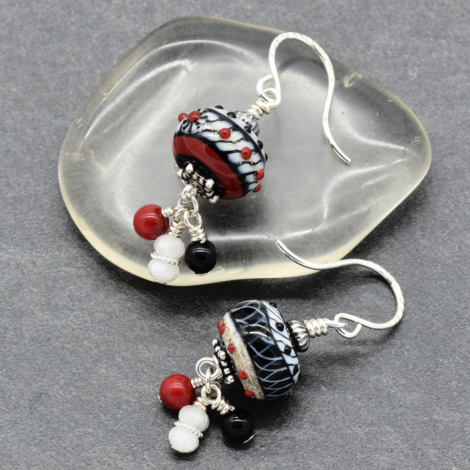 A pair of handmade lampwork dangle earrings displayed on a clear, organic-shaped glass dish over a dark gray textured background. The earrings feature a black and white patterned glass bead with red and black details, silver wire wrapping, and dangling red, white, and black beads - made with love in Colorado by Ruby Acorn.
