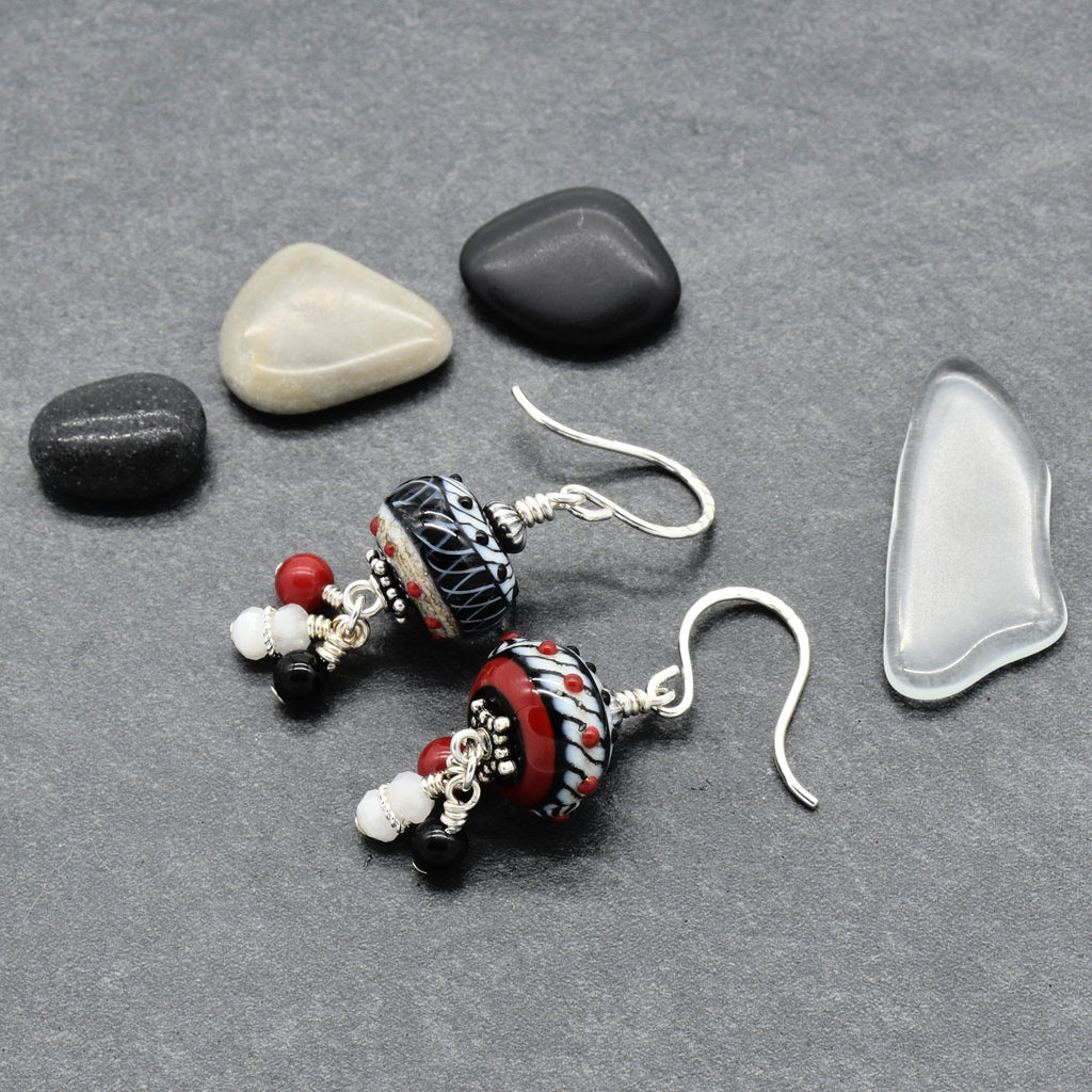 A pair of handmade beaded dangle earrings with silver fishhook wires, featuring patterned glass beads in black, white, red, and beige, displayed on a gray textured surface with decorative smooth stones - made with love in Colorado by Ruby Acorn.