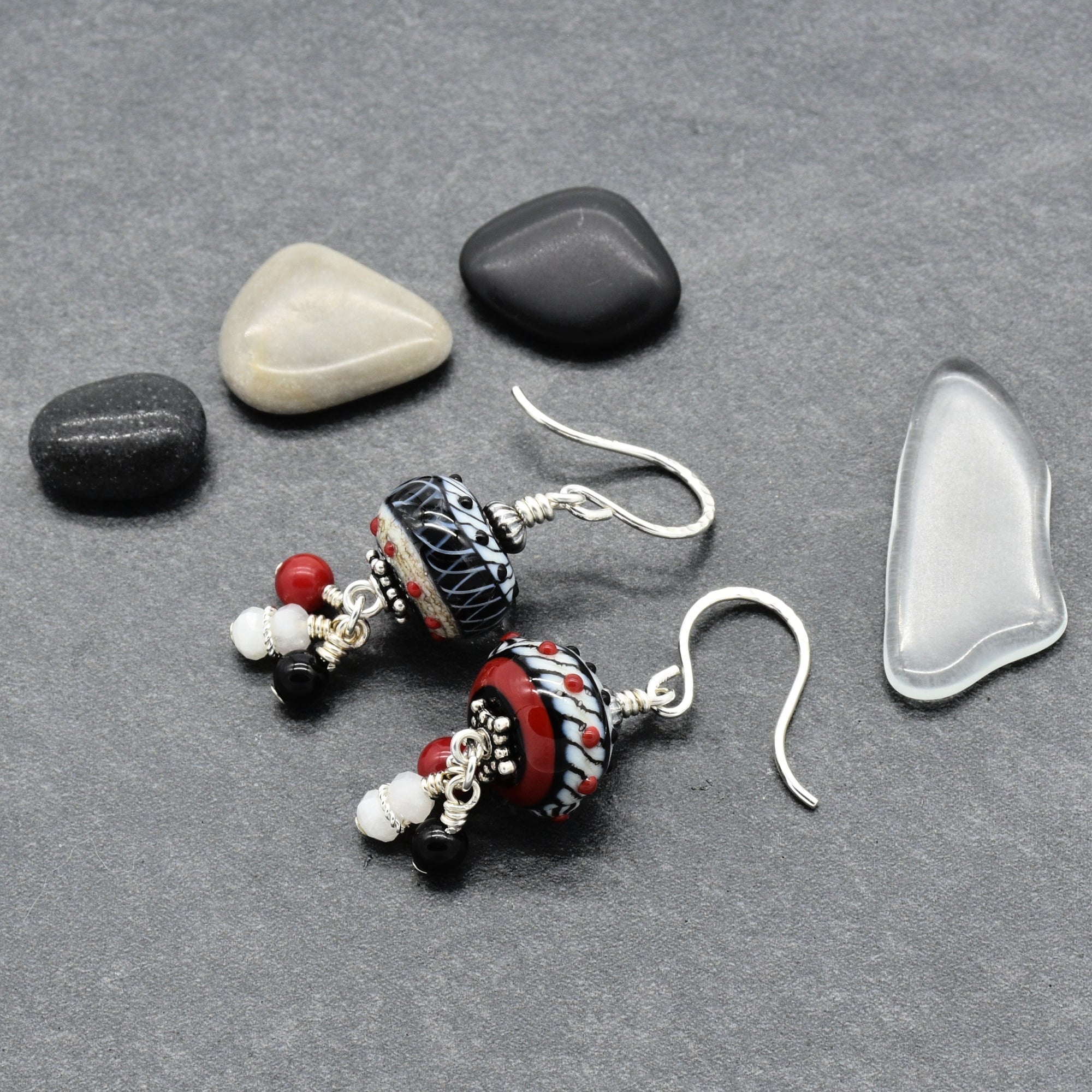 A pair of handmade beaded dangle earrings with silver fishhook wires, featuring patterned glass beads in black, white, red, and beige, displayed on a gray textured surface with decorative smooth stones - made with love in Colorado by Ruby Acorn.
