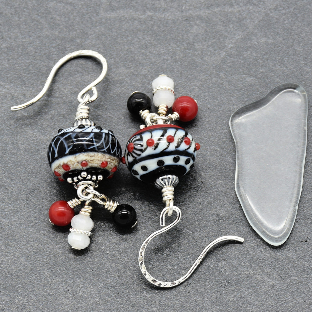 A pair of handmade glass bead drop earrings with silver fishhook wires, displayed next to a clear glass pebble on a textured gray background. The beads feature black and white patterns with red and white accents - made with love in Colorado by Ruby Acorn.