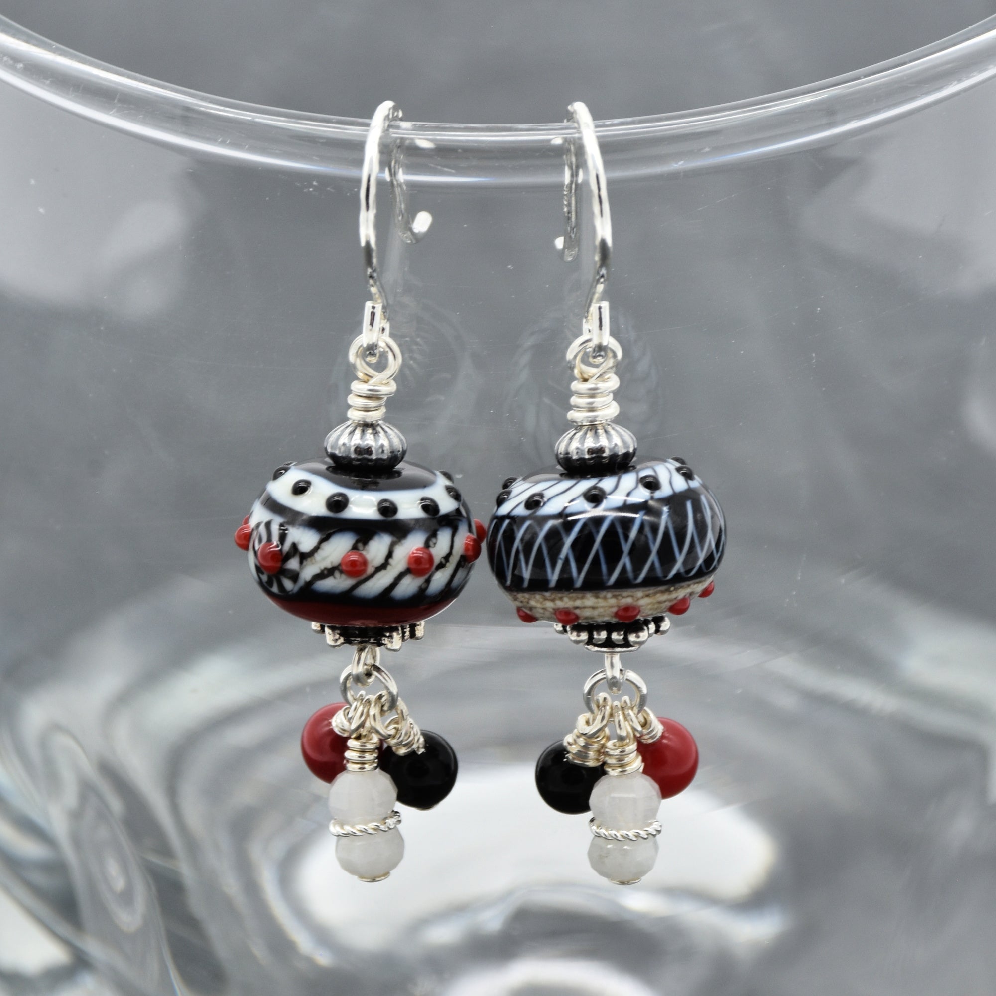 A pair of handmade lampwork earrings with black, white, and red patterned beads, hanging from silver fishhook wires against a clear glass background - made with love in Colorado by Ruby Acorn.
